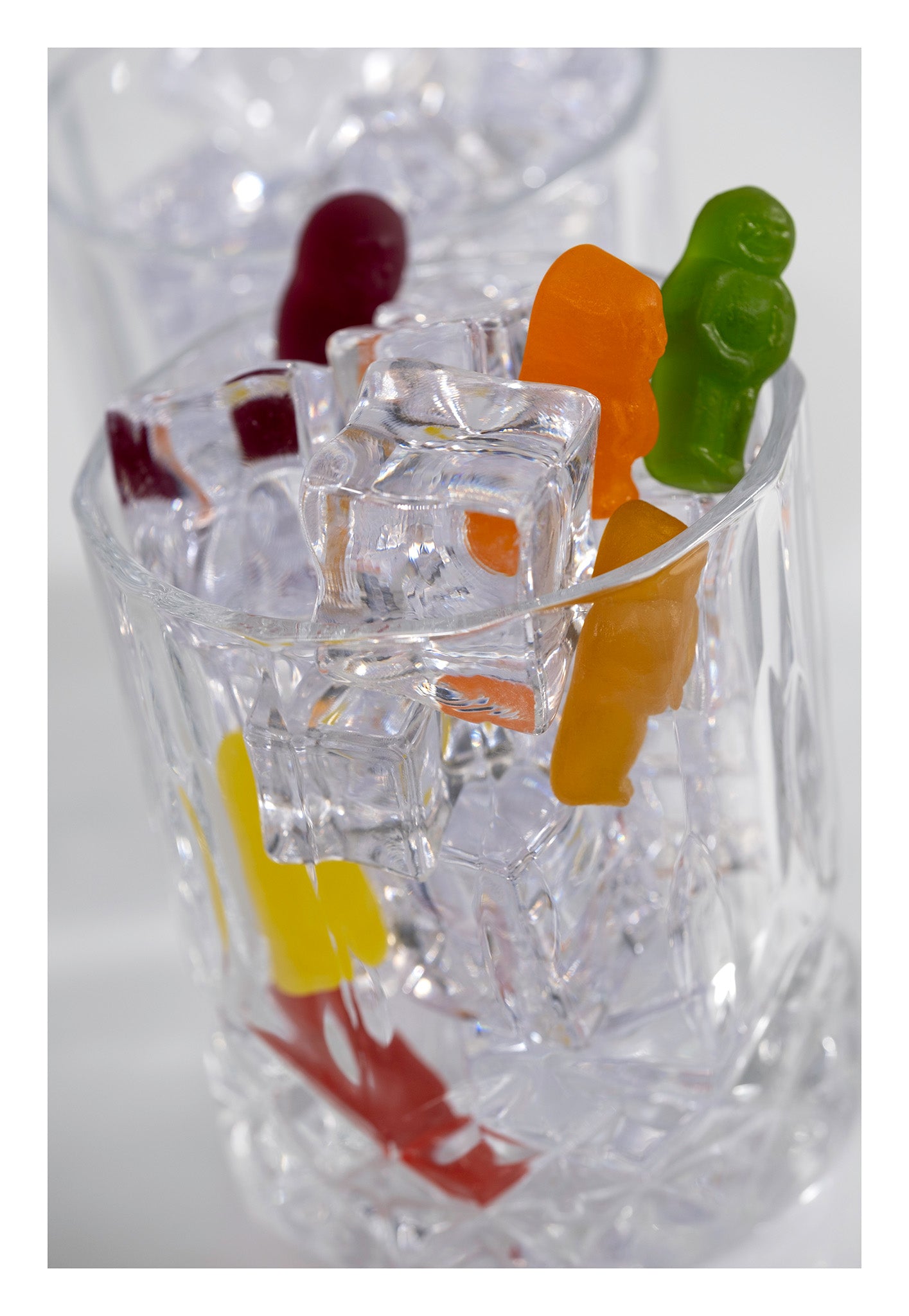 Colourful gummy figures arranged among transparent ice cubes inside a clear glass, photographed against a pale background.