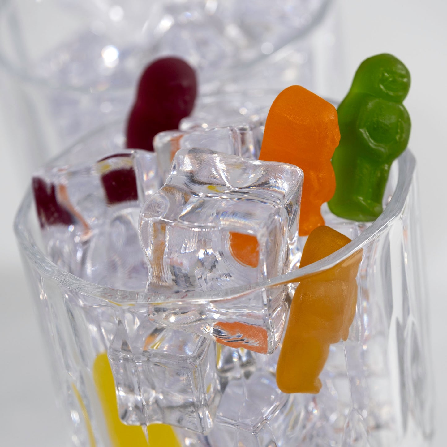 Close-up of orange and green gummy figures perched among transparent ice cubes in a clear glass, with a red figure softly focused behind.