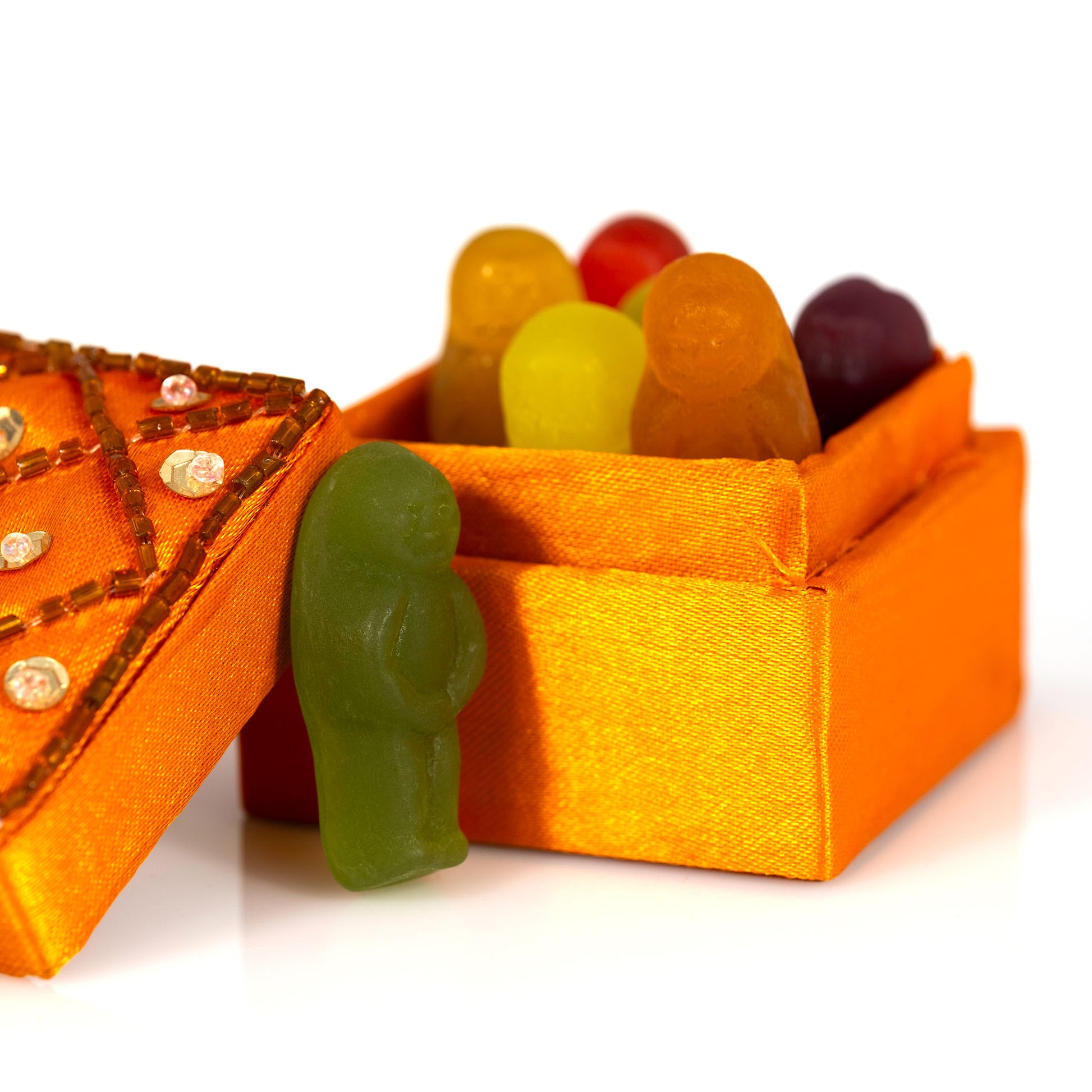 Close-up of a green gummy figure standing outside an orange fabric-covered box containing a group of colourful gummy figures.