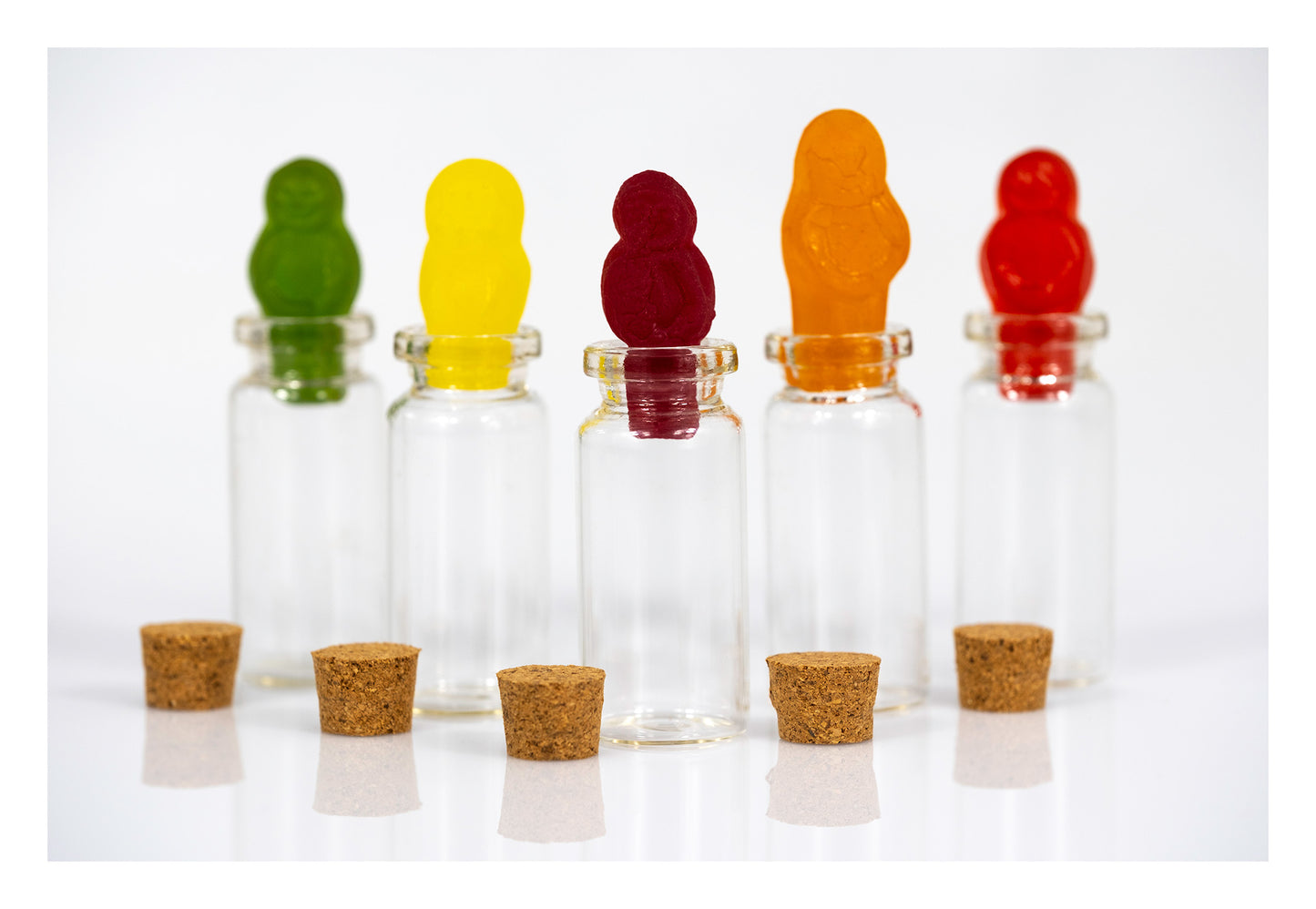 Five colourful gummy figures positioned in the openings of transparent glass bottles with five small cork stoppers arranged in front.