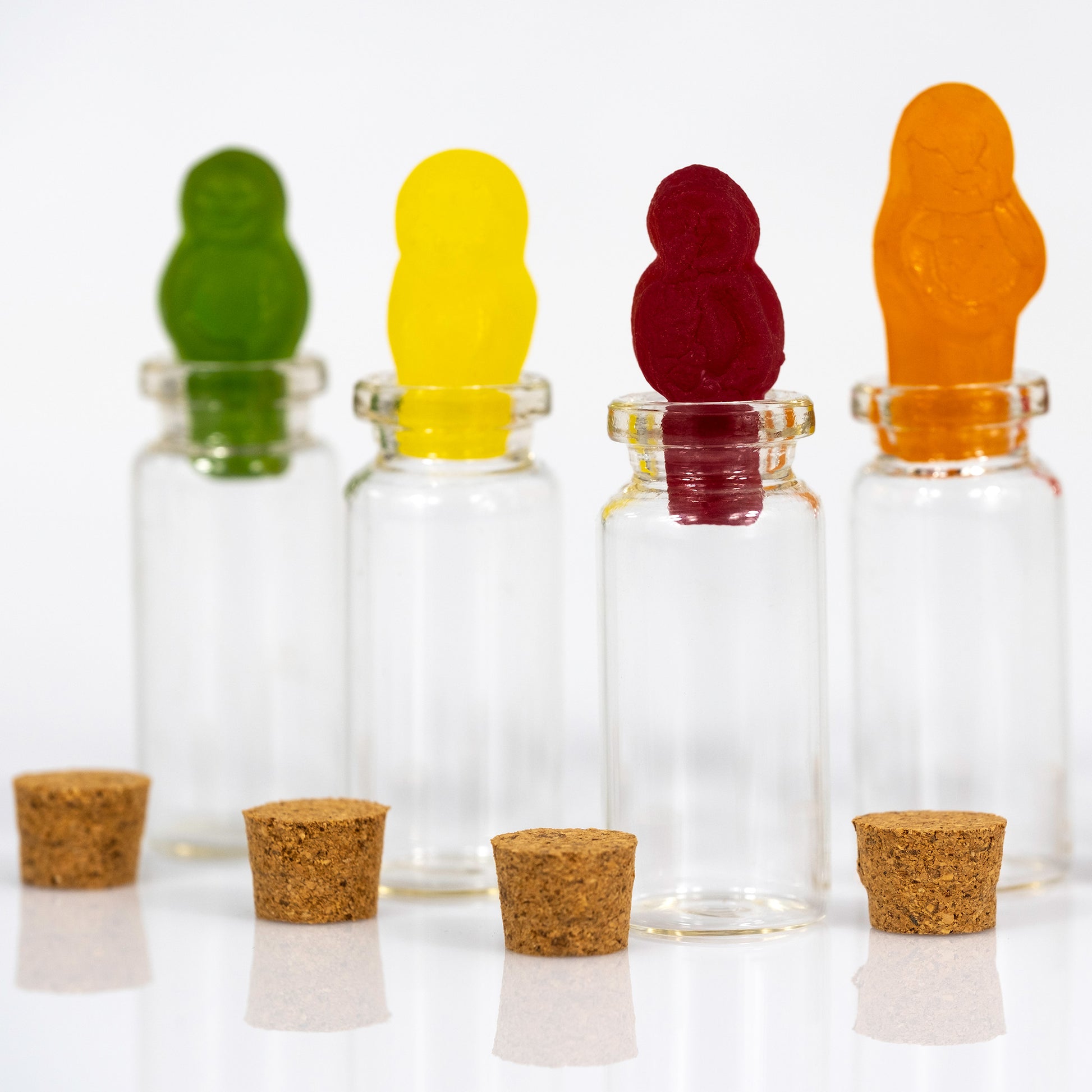 Close view of colourful gummy figures standing in clear glass bottles, with cork stoppers reflected on the white surface below.