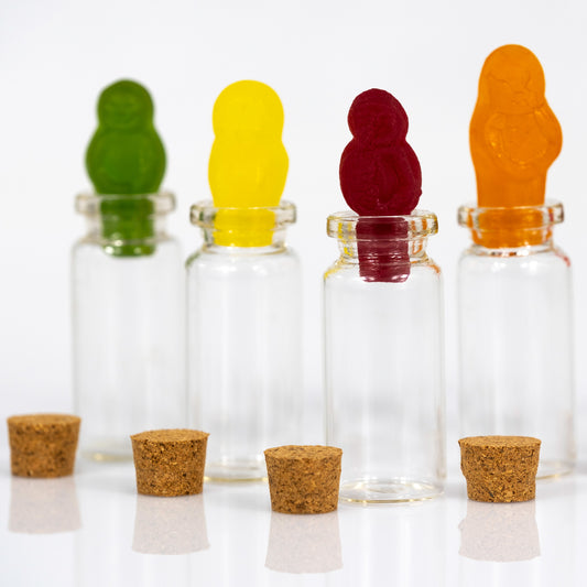 Close view of colourful gummy figures standing in clear glass bottles, with cork stoppers reflected on the white surface below.