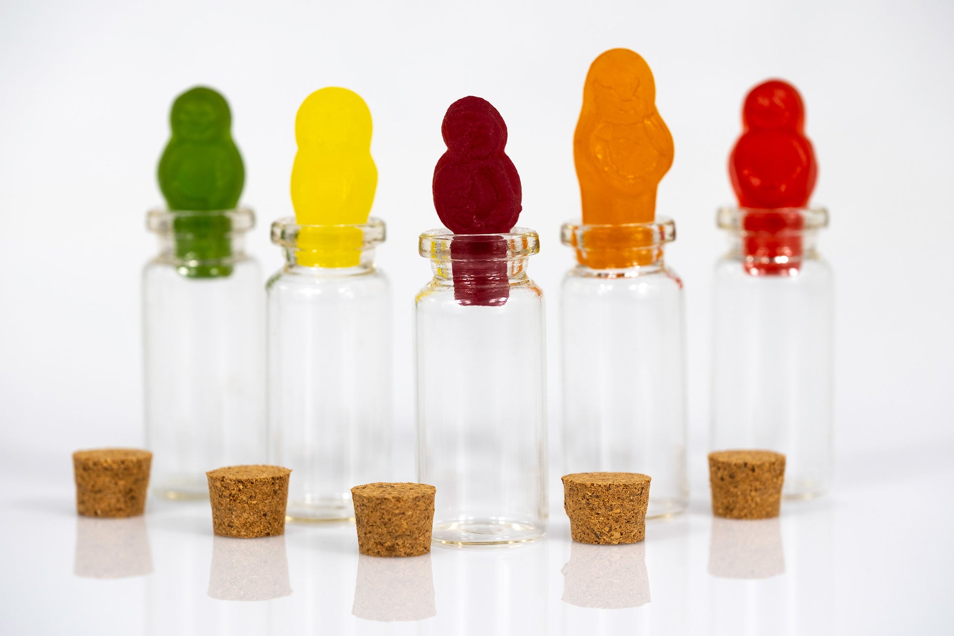 Five green, yellow, burgundy, orange and red gummy figures arranged across transparent glass bottles against a white background.