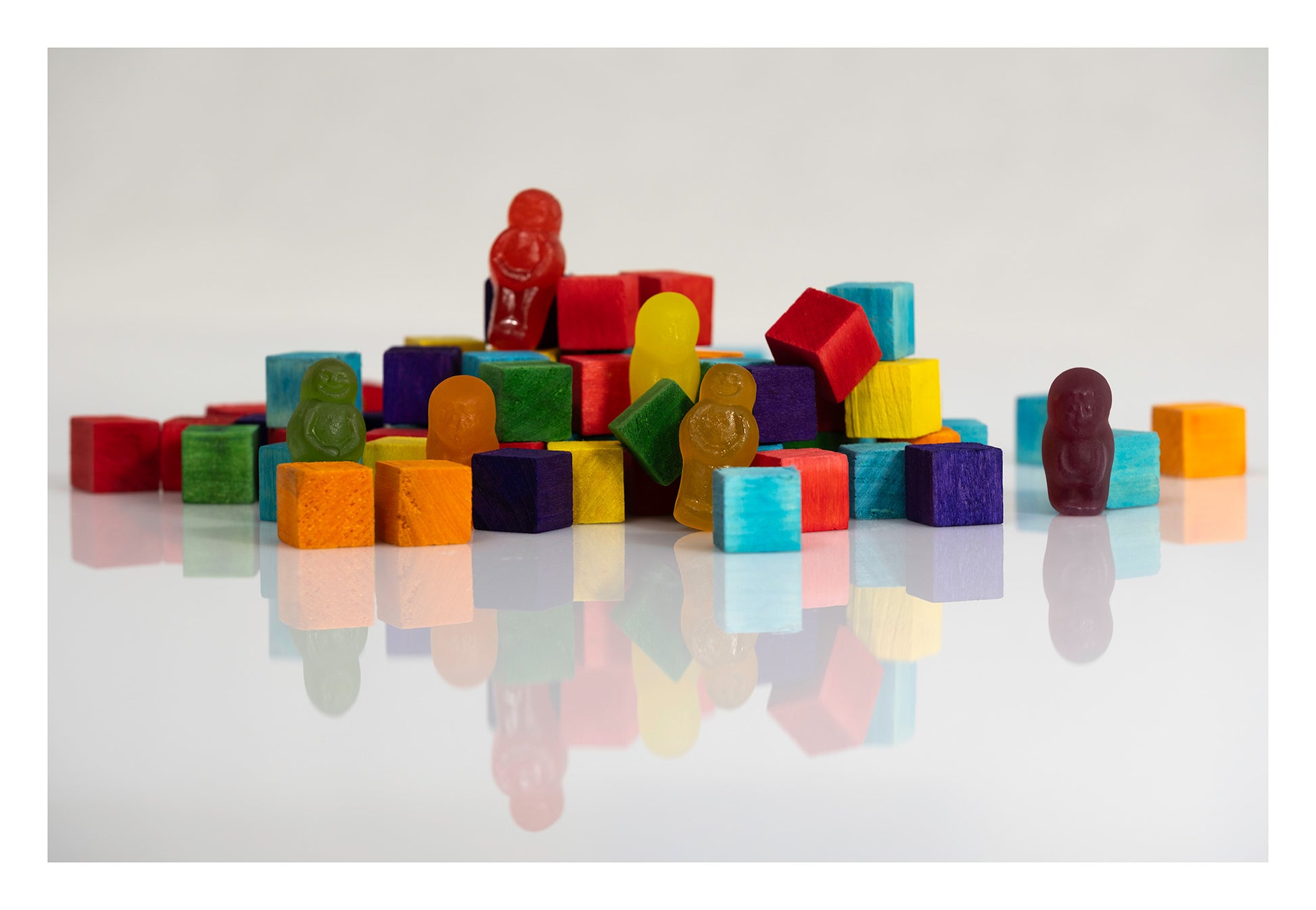 Colourful wooden blocks and gummy figures arranged on a reflective white surface, with one purple figure standing apart on the right.