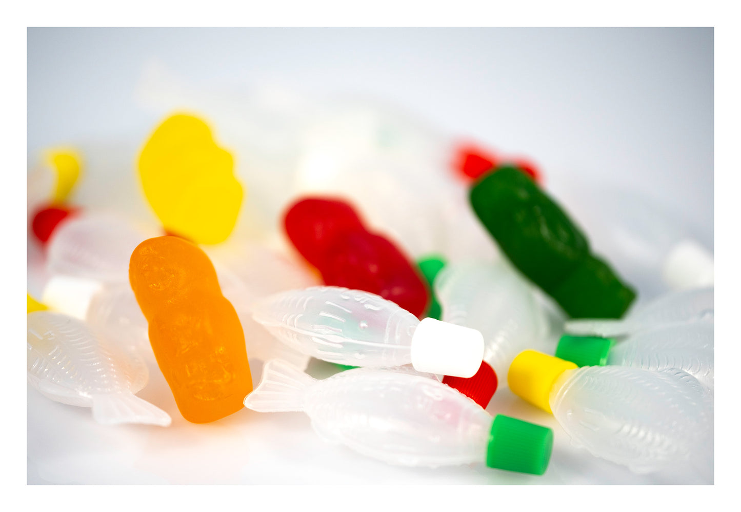 Orange, red, green and yellow gummy figures surrounded by translucent fish-shaped objects against a bright white background.