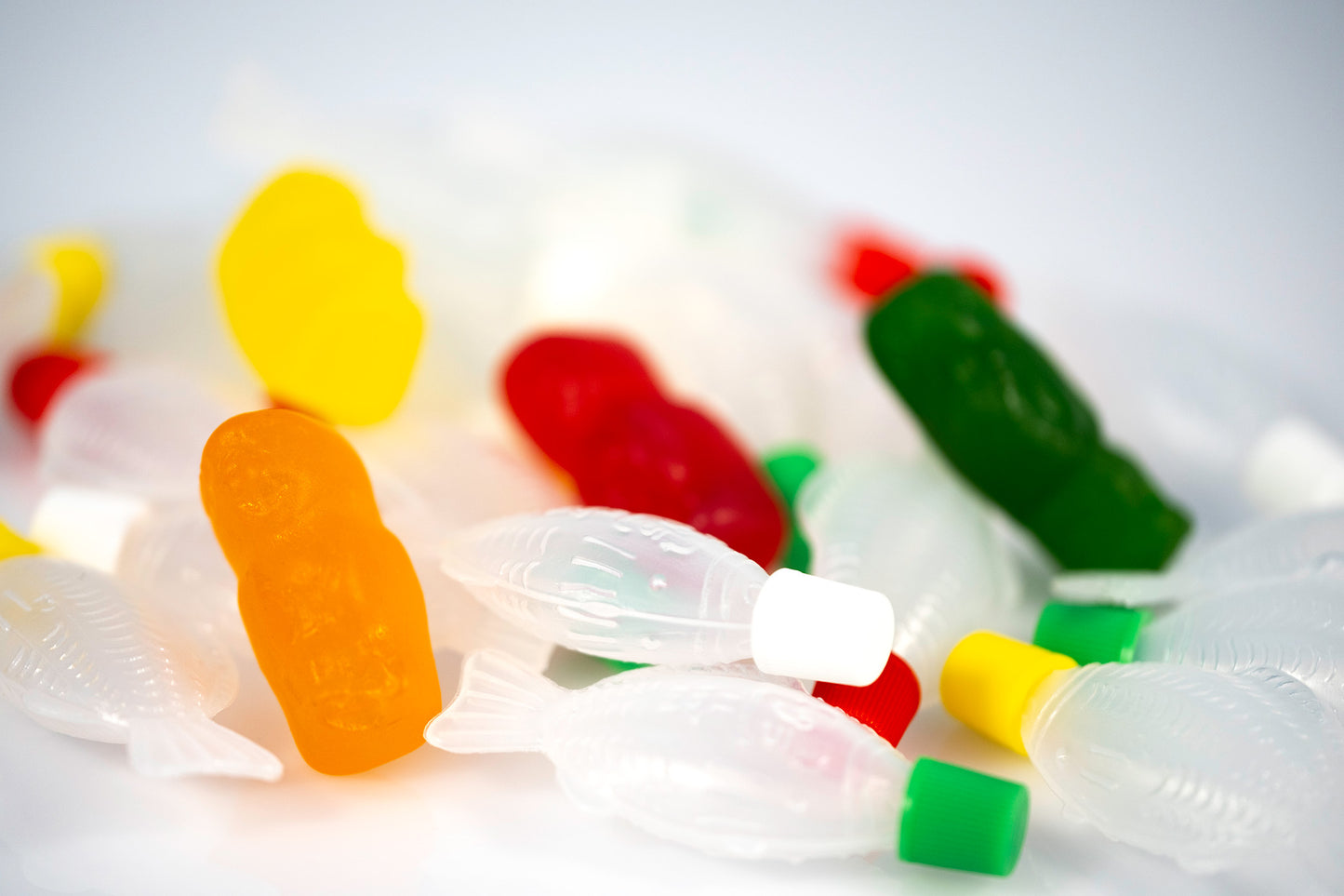 Conceptual still life of colourful gummy figures scattered among numerous translucent fish-shaped objects on a white surface.