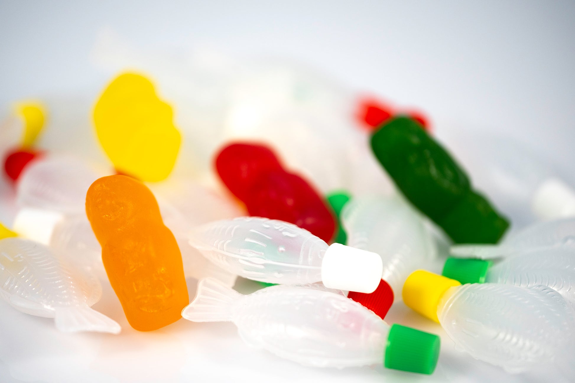 Conceptual still life of colourful gummy figures scattered among numerous translucent fish-shaped objects on a white surface.