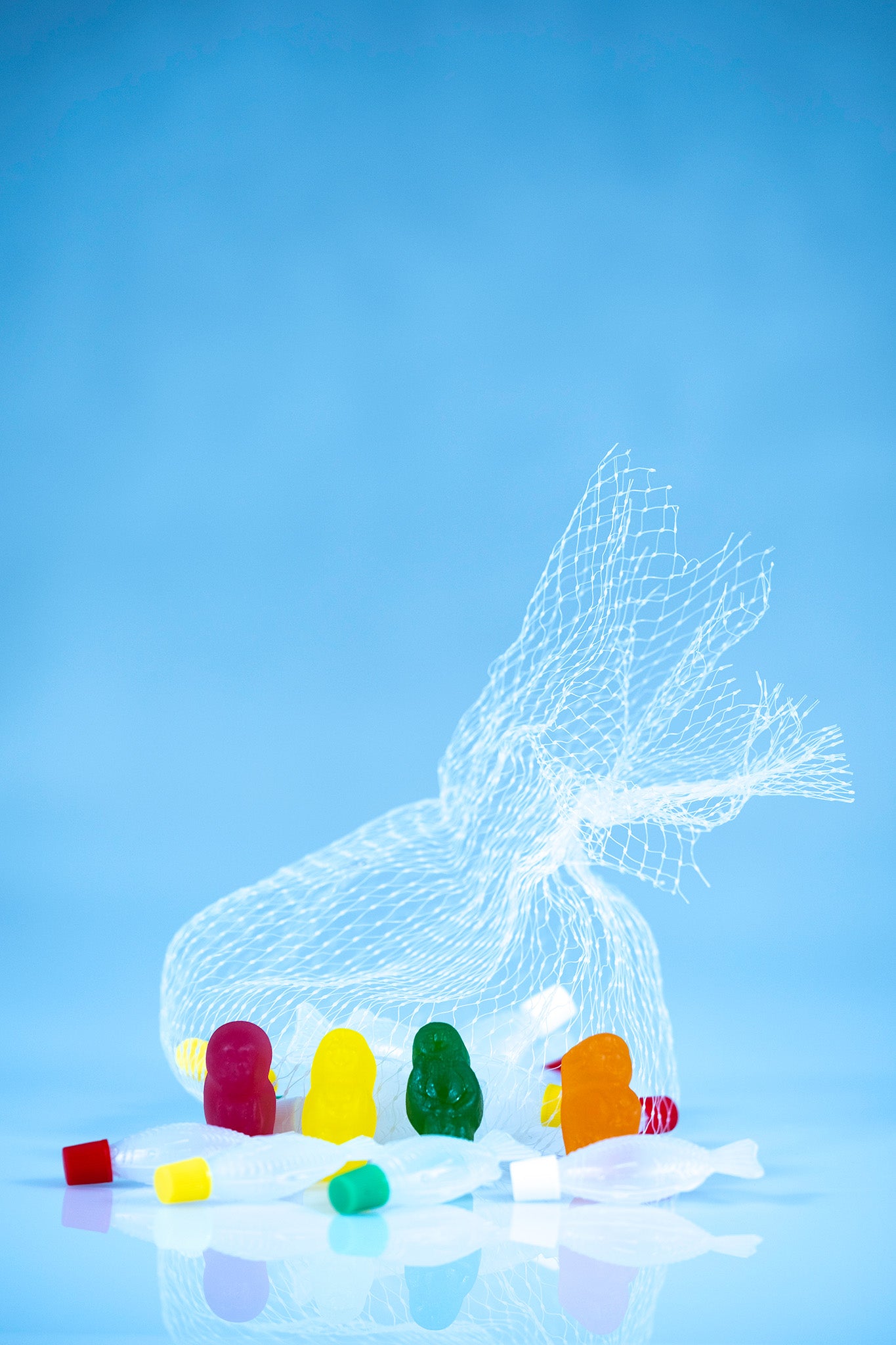 Portrait conceptual still life of four colourful gummy figures and scattered fish beneath an oversized white net on a blue background.