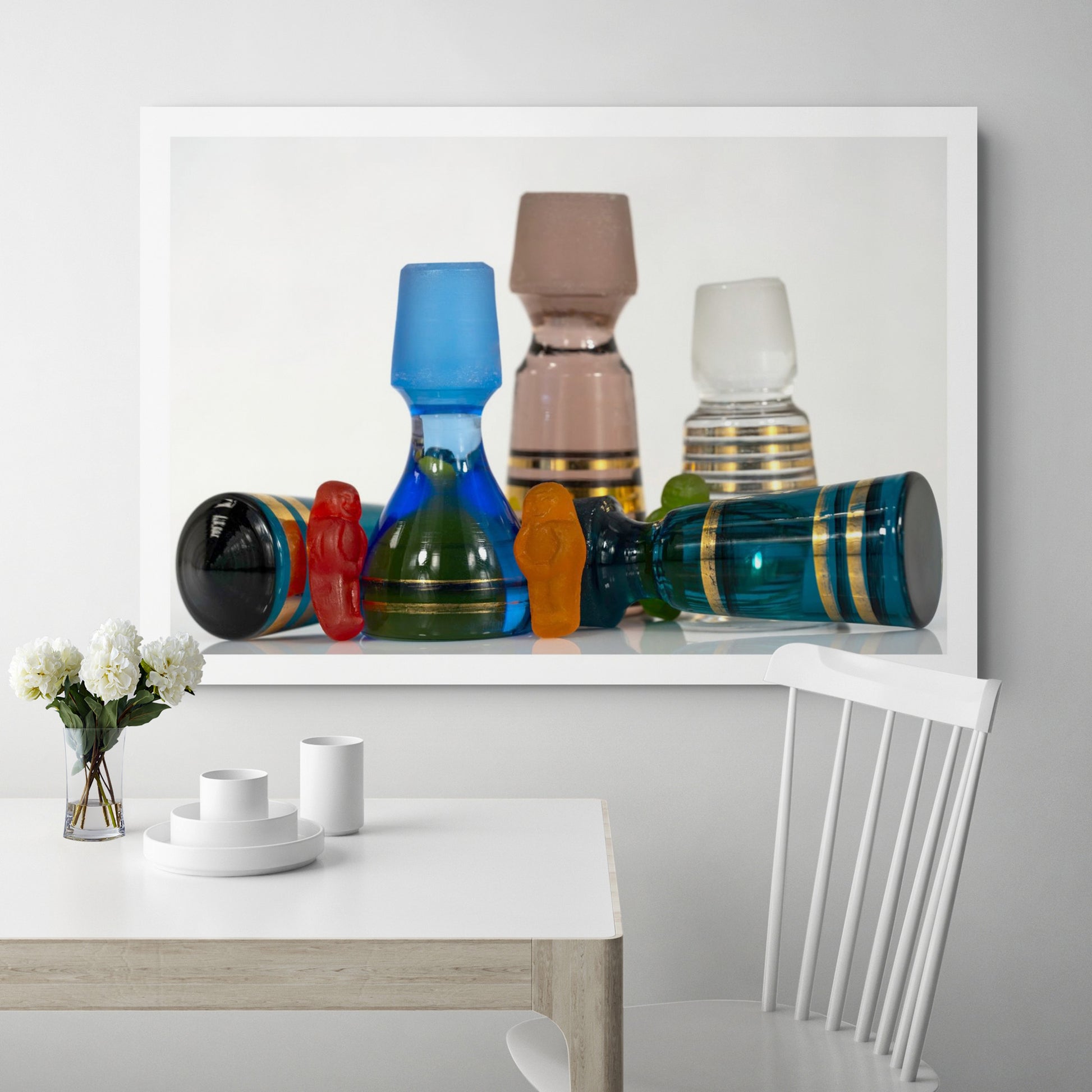 Large photographic artwork featuring colourful gummy figures among vintage blue, pink and clear glass stoppers, displayed above a contemporary dining table.