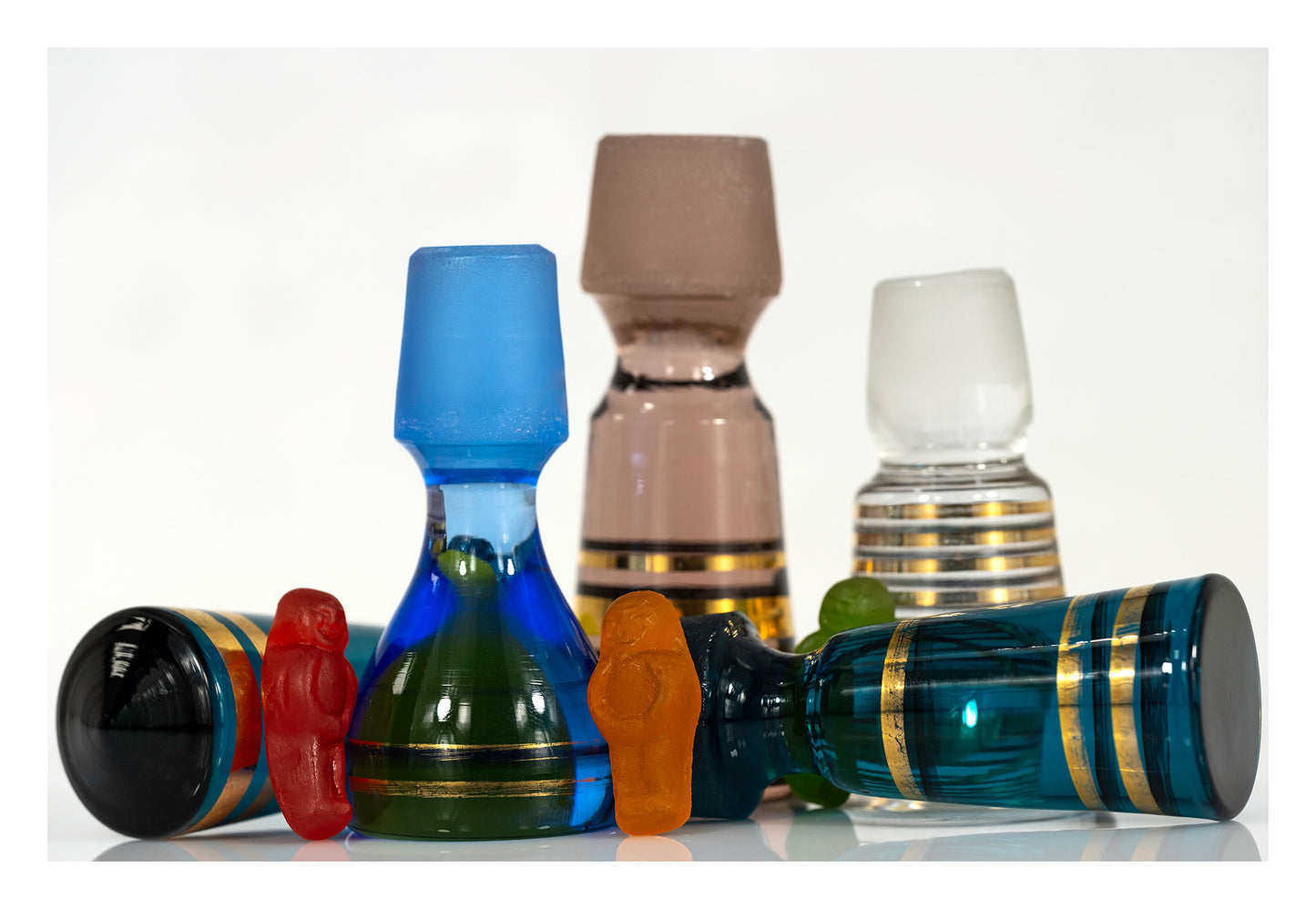 Red, orange and green gummy figures arranged among upright and fallen vintage glass stoppers on a white reflective surface.