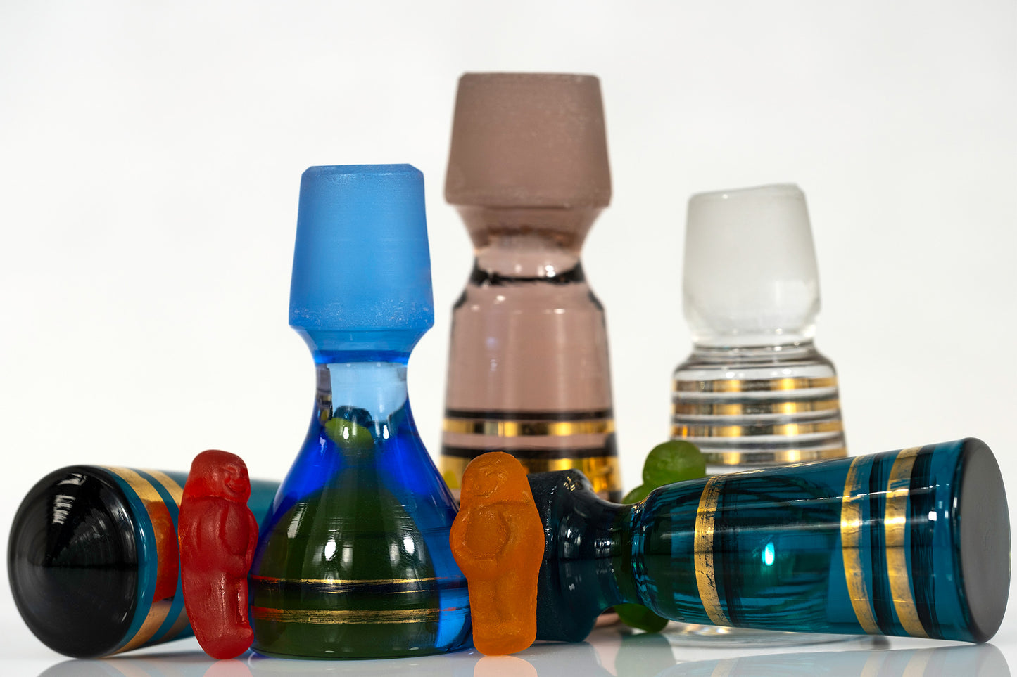 Colourful gummy figures standing among large decorative vintage glass stoppers, with blue and teal stoppers lying horizontally in the foreground.