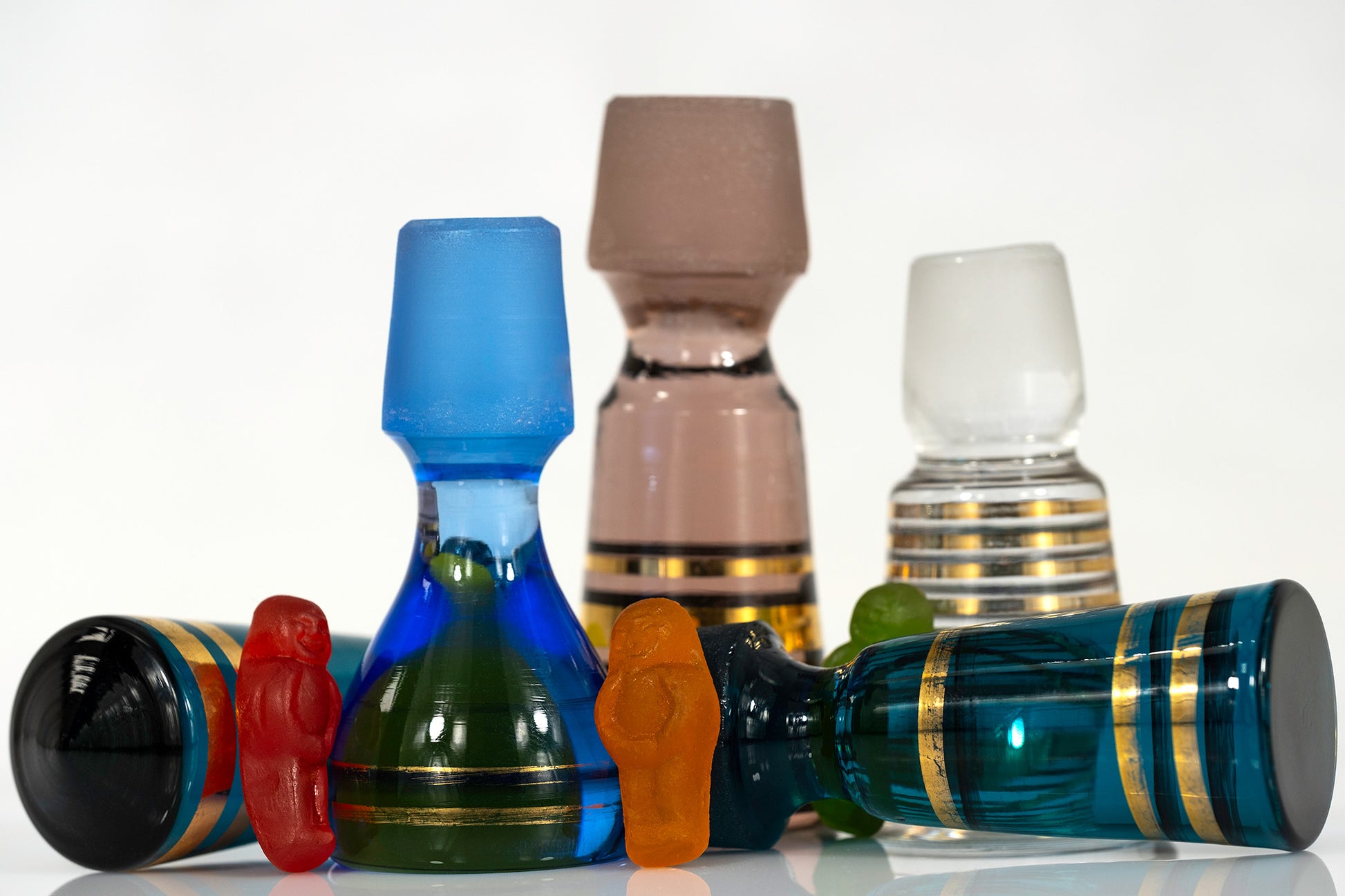 Colourful gummy figures standing among large decorative vintage glass stoppers, with blue and teal stoppers lying horizontally in the foreground.