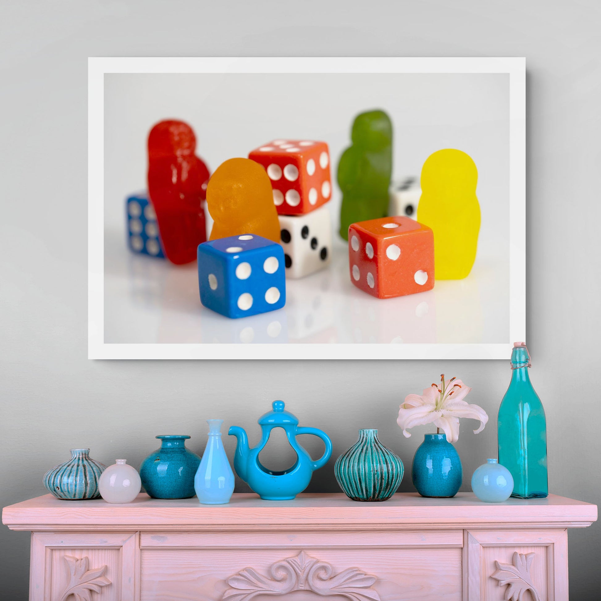 Colourful photographic artwork of gummy figures and red, blue and white dice displayed above a pale pink cabinet with blue decorative ceramics.