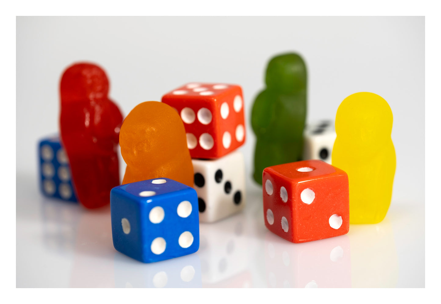 Colourful gummy figures arranged among red, blue and white dice on a reflective white surface.
