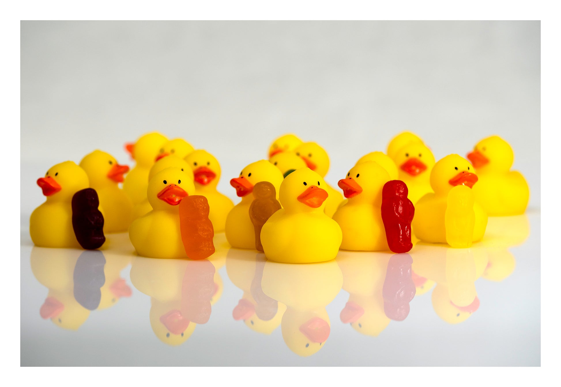 Yellow rubber ducks gathered with small colourful figures on a reflective white surface.