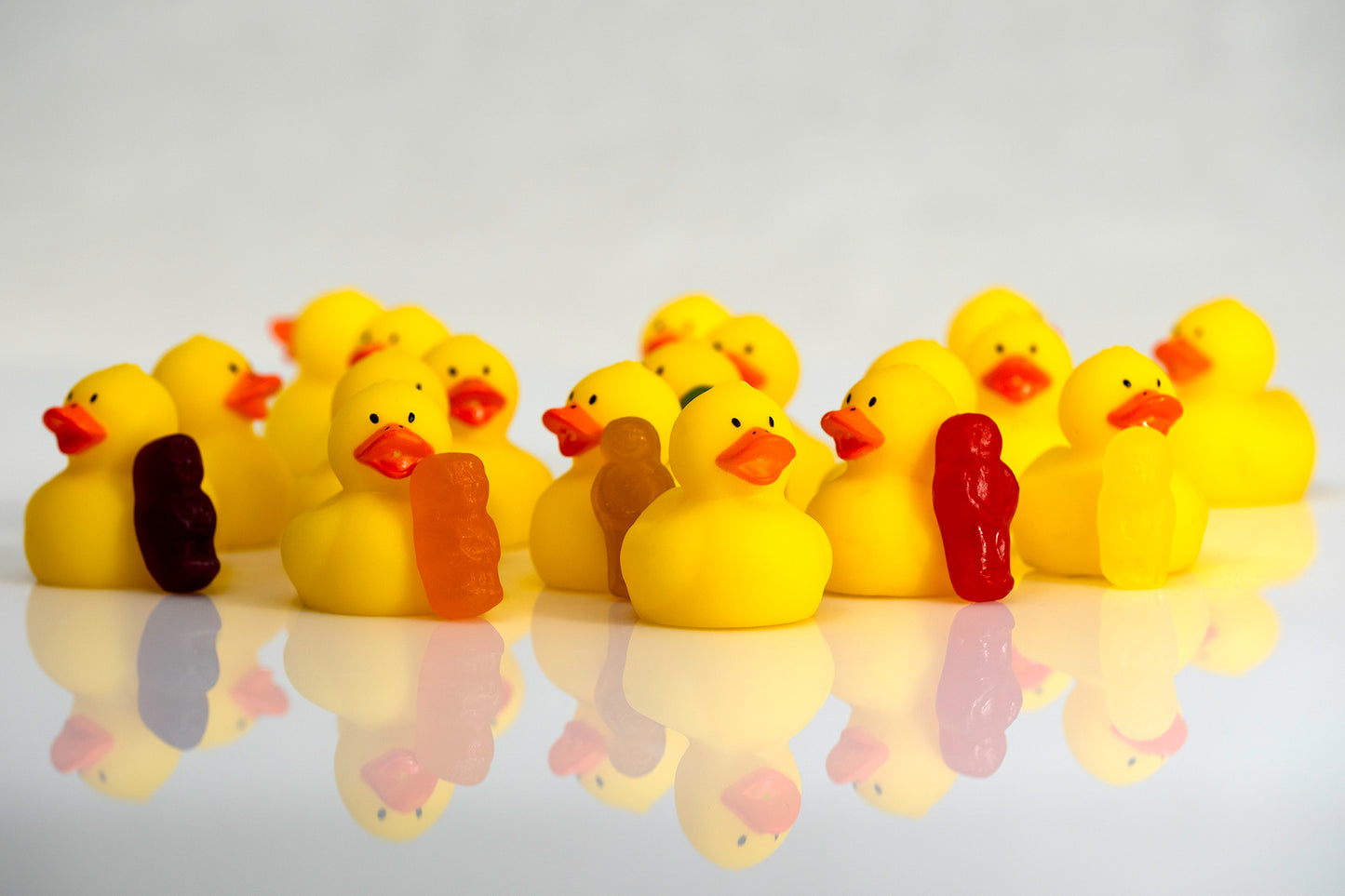 Crowd of bright yellow rubber ducks interspersed with small colourful figures on a reflective surface.