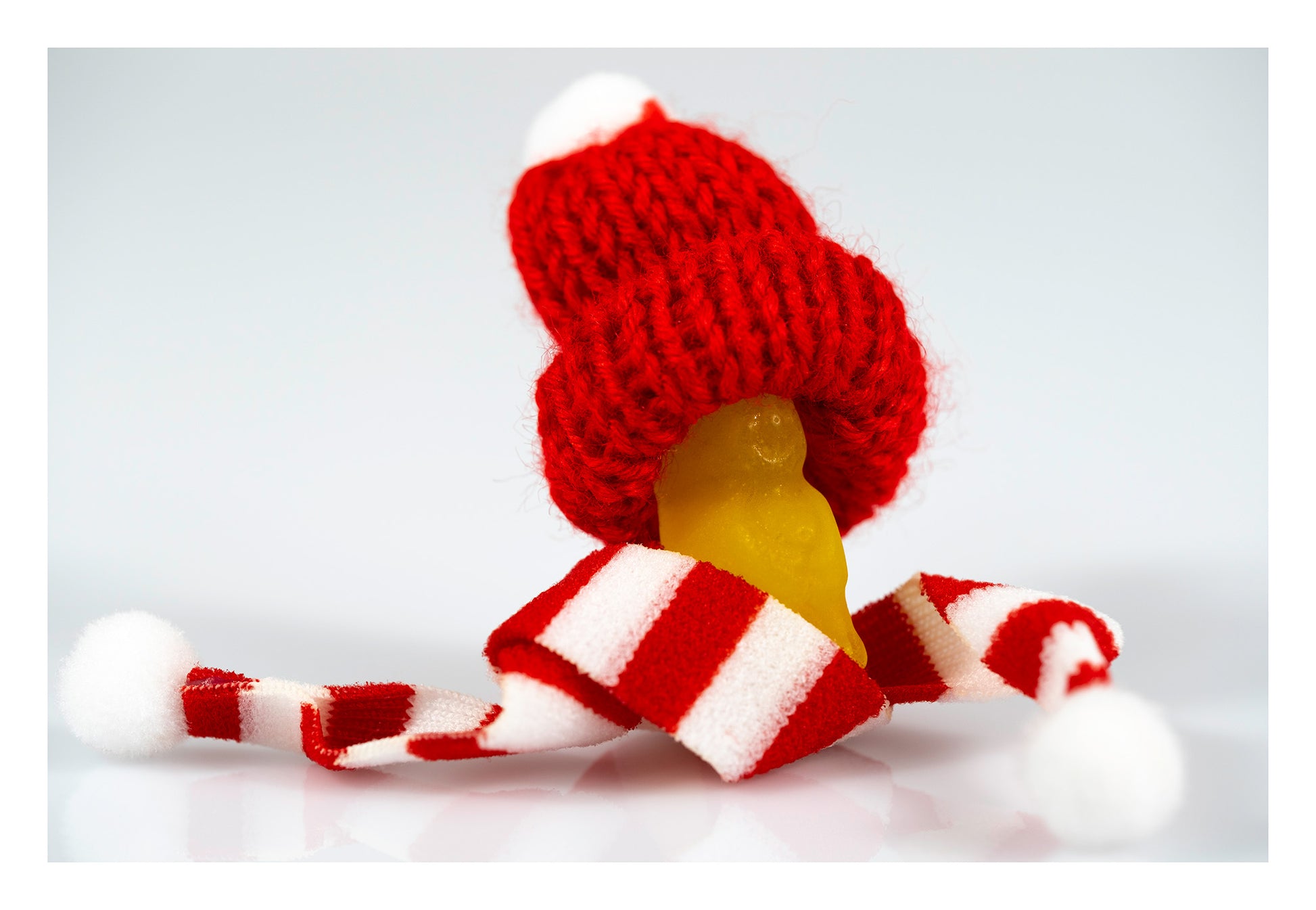 Tiny yellow figure wearing an oversized red knitted Santa hat and red-and-white striped scarf against a pale background.