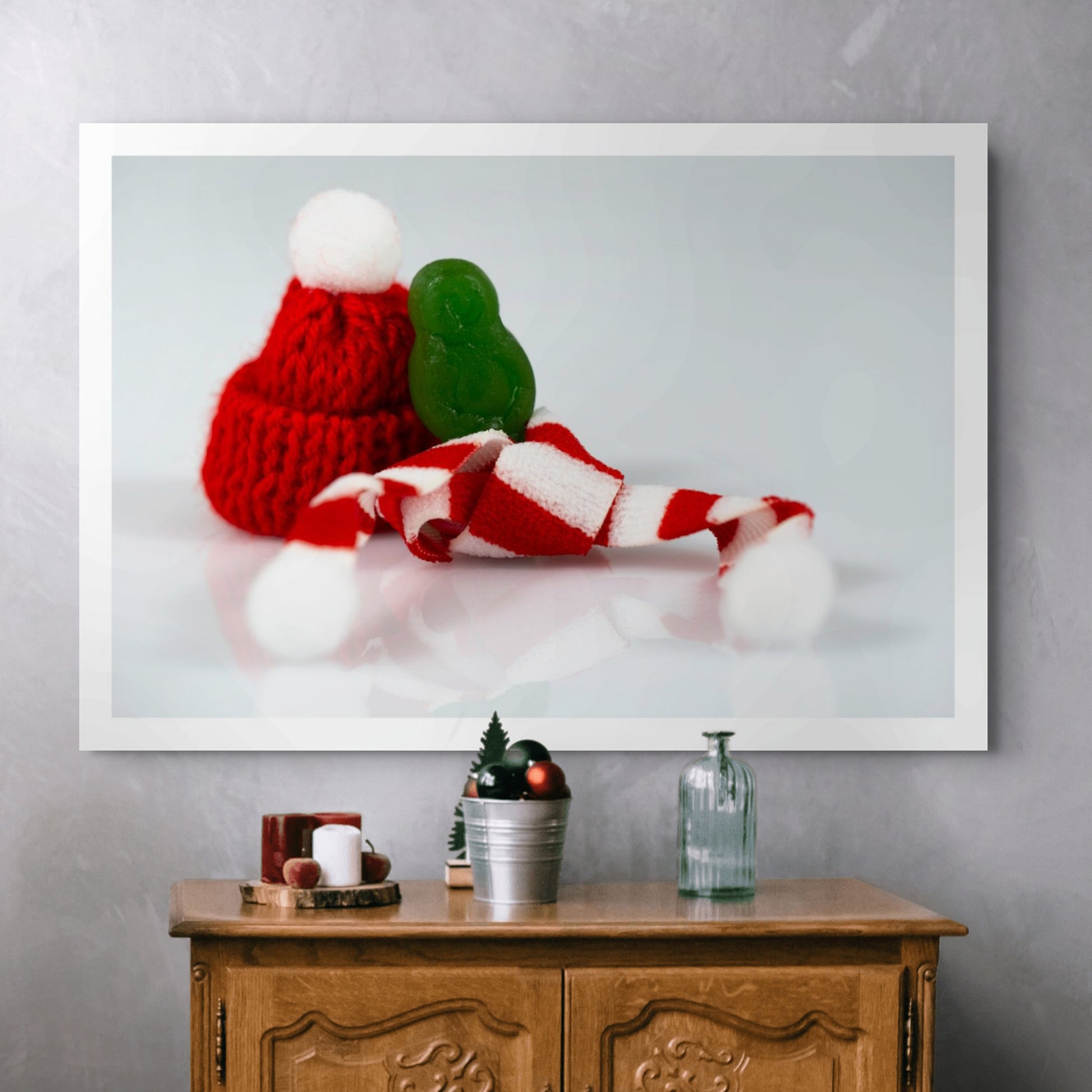 Santa Babies photographic artwork showing a tiny green figure beside a red Santa hat and striped scarf displayed above a wooden cabinet.