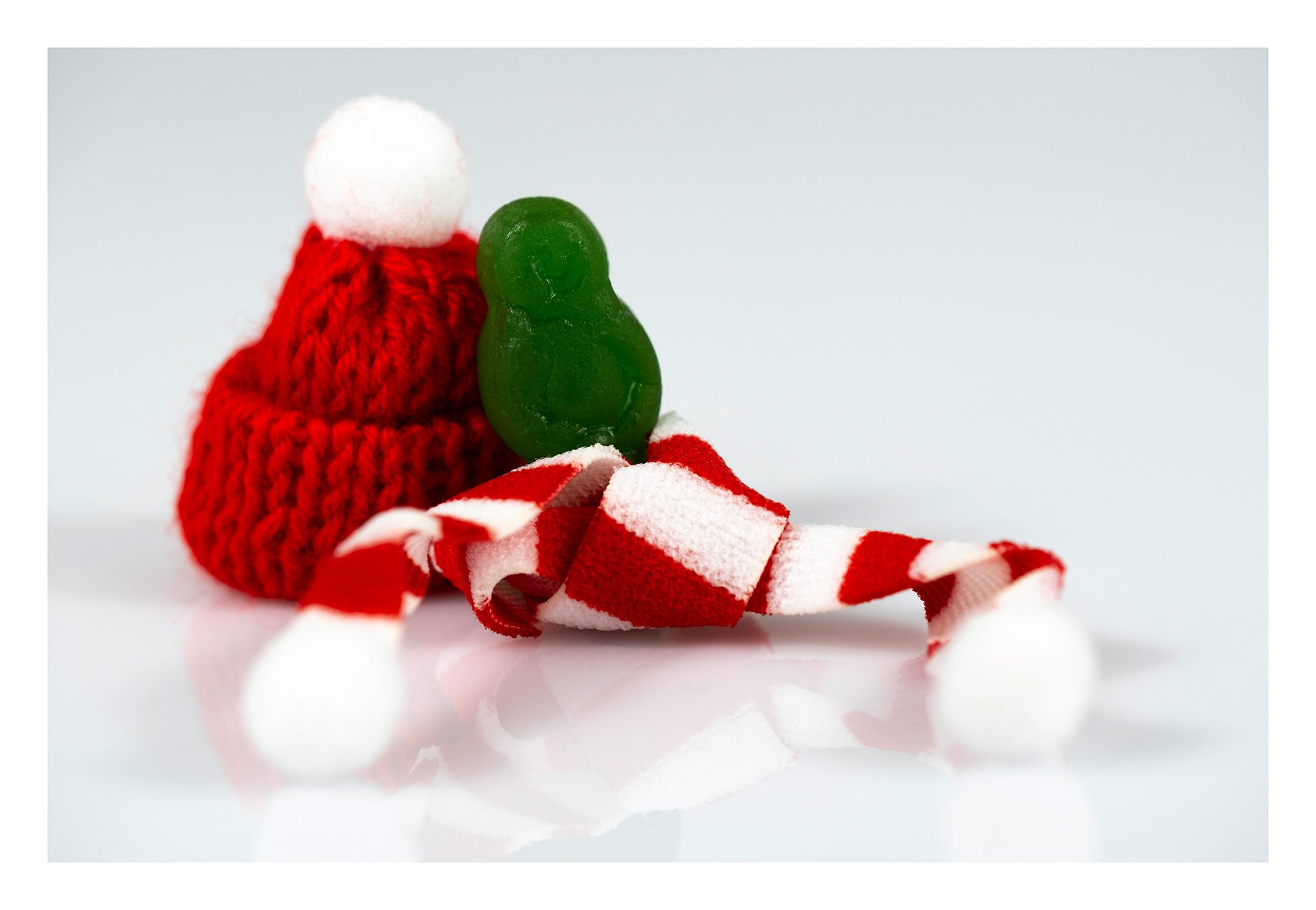 Tiny green figure sitting beside a red knitted Santa hat with a red-and-white striped scarf spread across a reflective surface.
