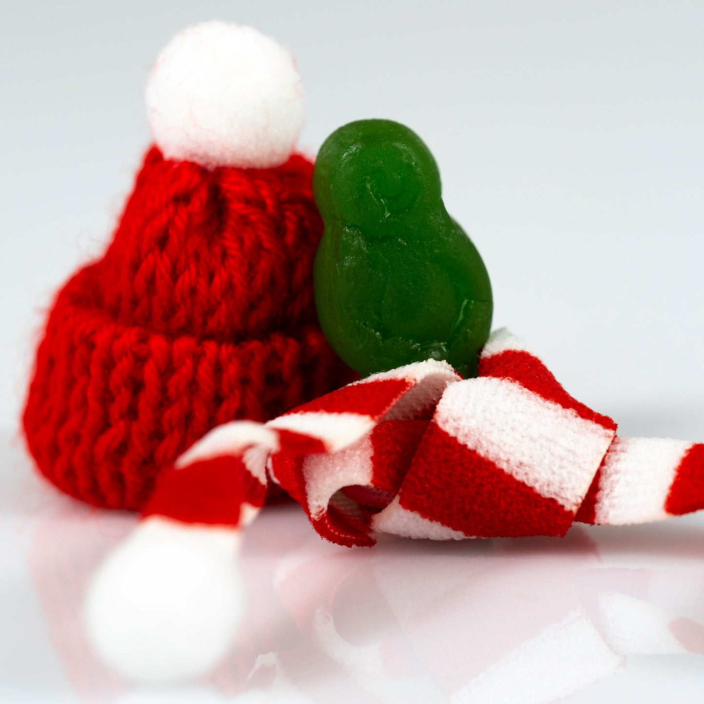 Close-up of a small green figure beside an oversized red knitted Santa hat and folded striped scarf.
