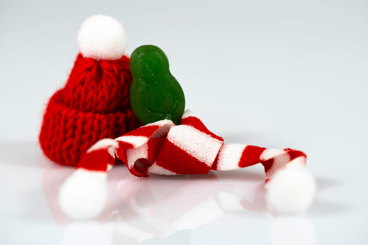 Green miniature figure with discarded red Santa hat, striped scarf and white pompoms against a pale background.
