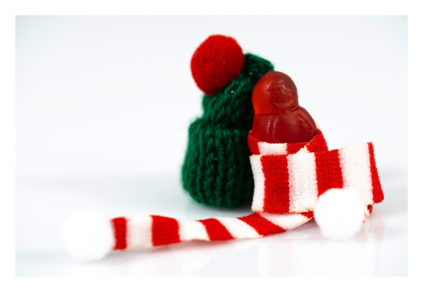 Tiny red figure wrapped in a red-and-white striped scarf beside a green knitted hat on a reflective white surface.