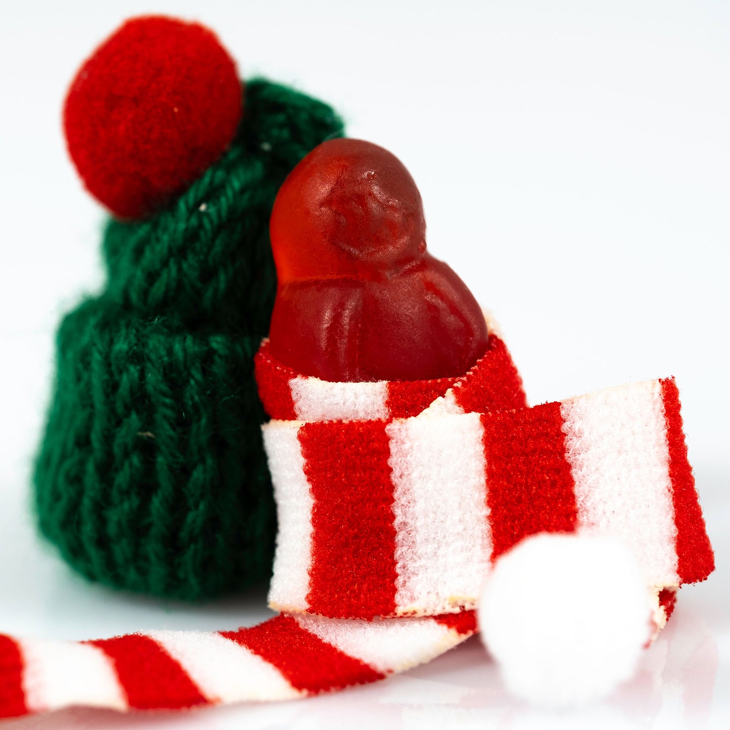 Close-up portrait of a miniature red figure emerging from a striped Christmas scarf with a green knitted hat behind.