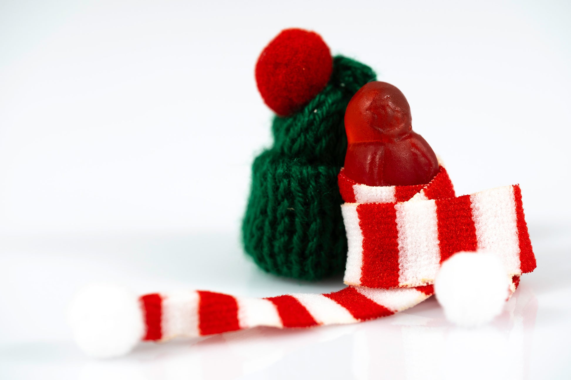 Red miniature figure wrapped in an oversized striped scarf beside a green knitted hat and white pompoms.