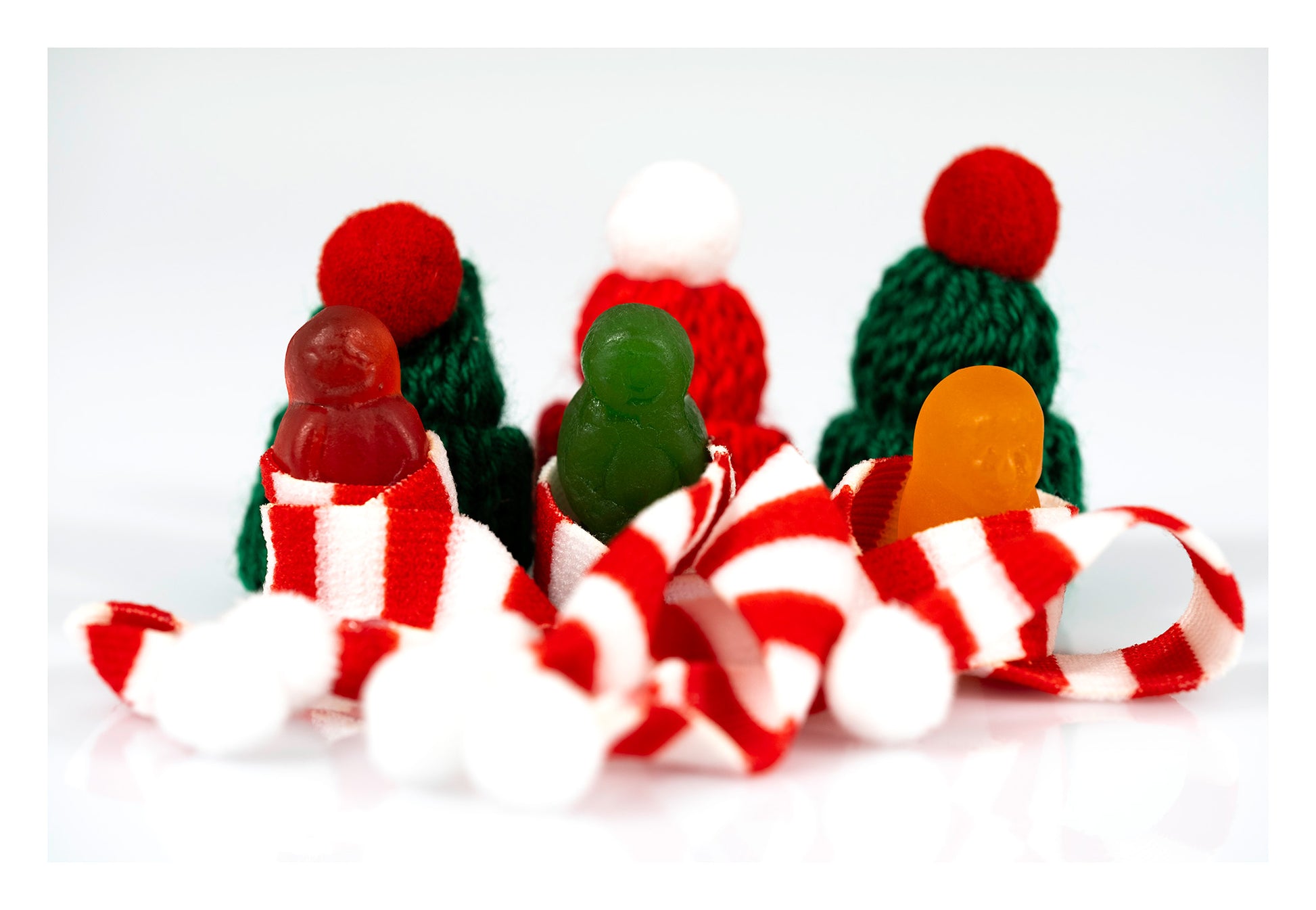 Three red, green and orange miniature figures wrapped in striped Christmas scarves with knitted hats behind them.