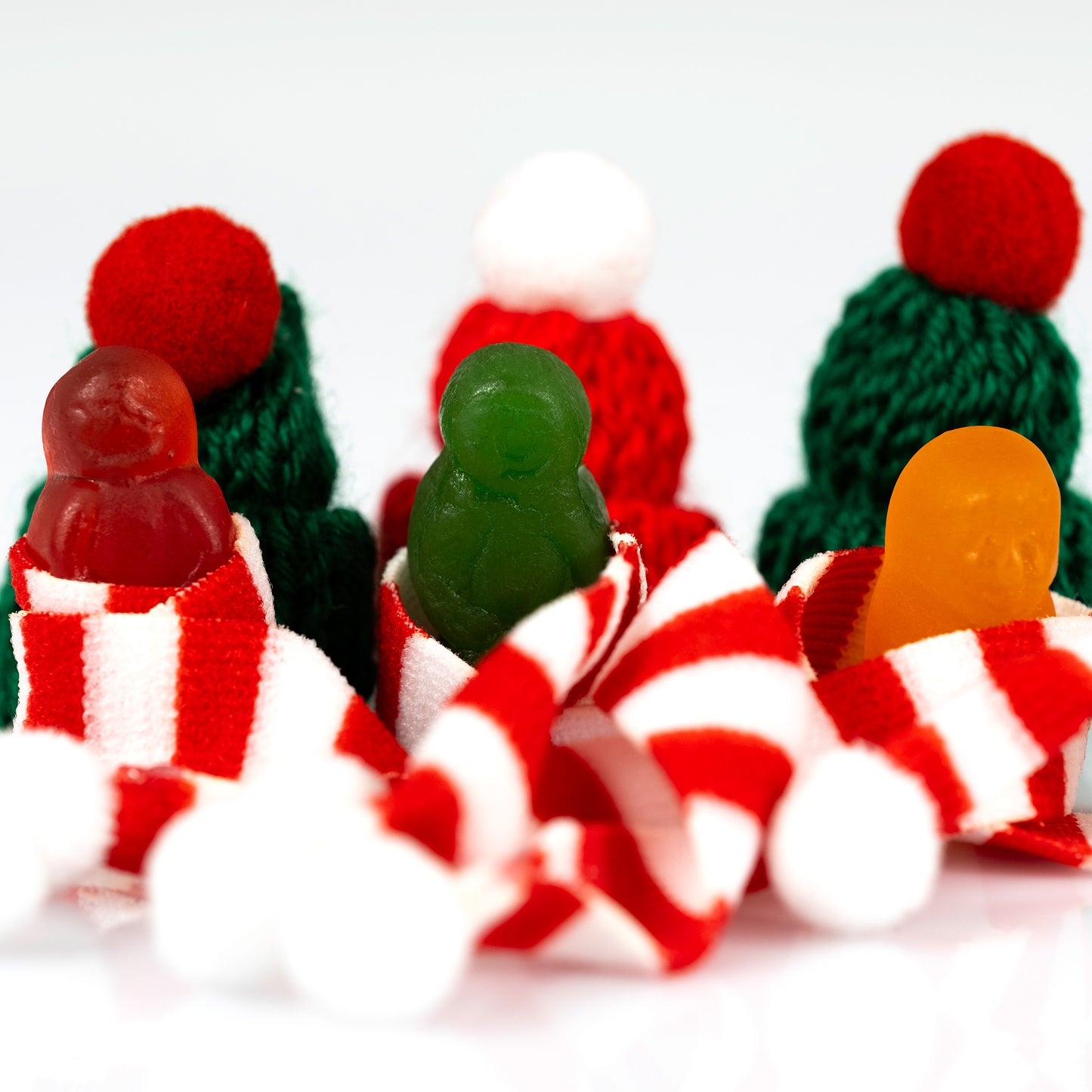 Close-up of three colourful miniature figures emerging from red-and-white striped scarves with knitted Christmas hats behind.