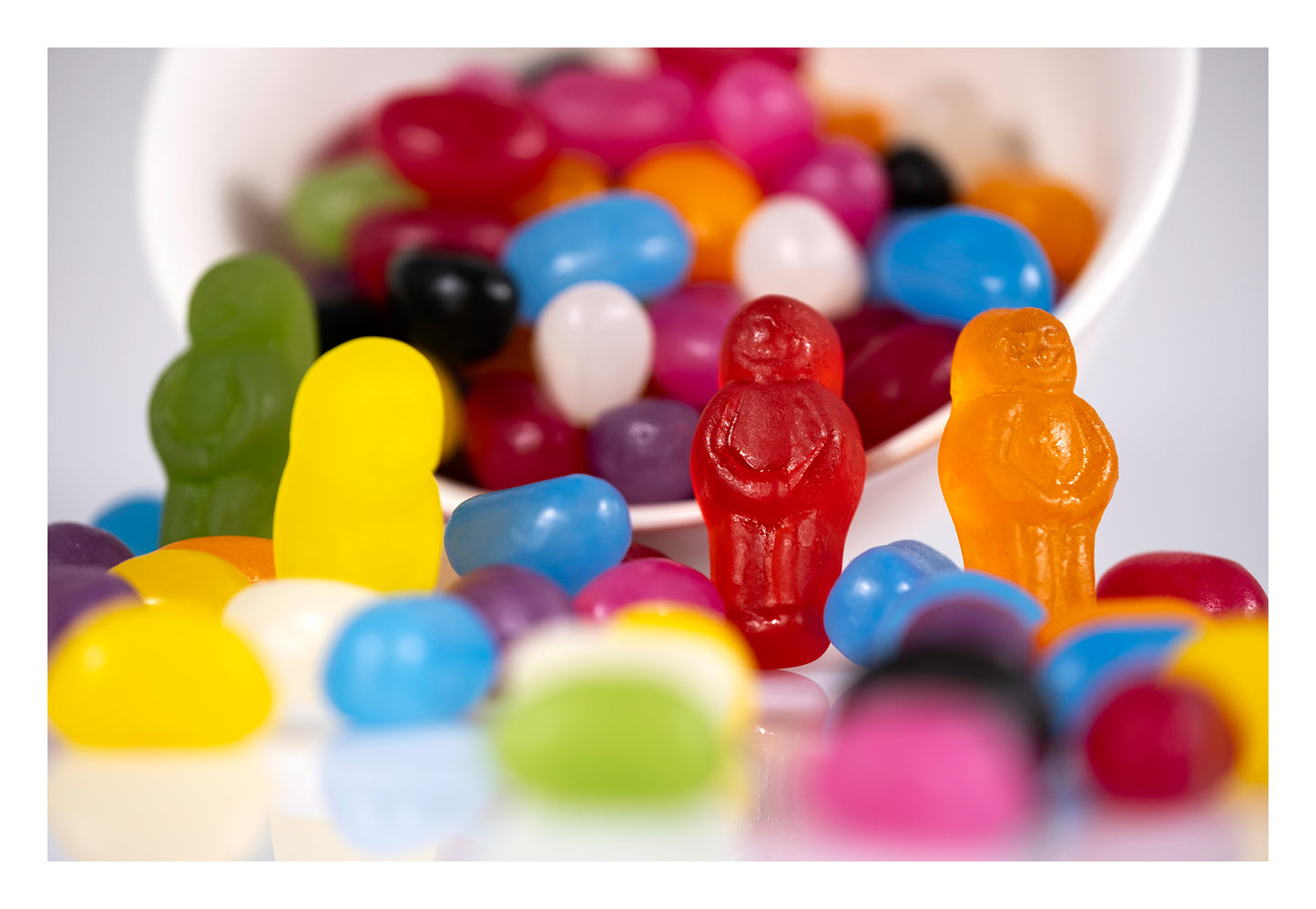 Four colourful jelly baby figures stand among scattered jelly beans spilling from a white bowl in Spill the Beans - 266468v1.