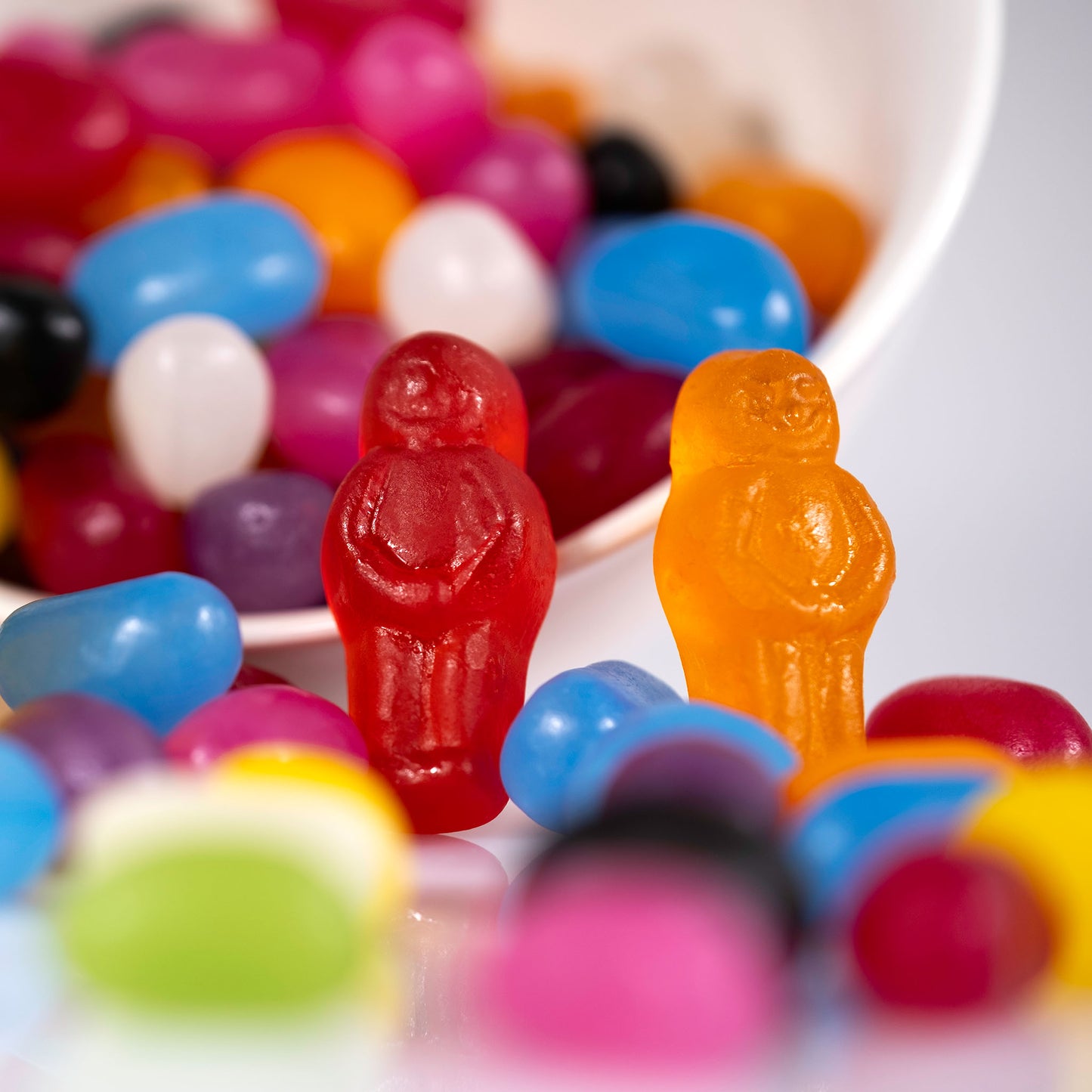 Red and orange jelly baby figures stand among brightly coloured jelly beans with a white bowl behind them in Spill the Beans - 266468v1.