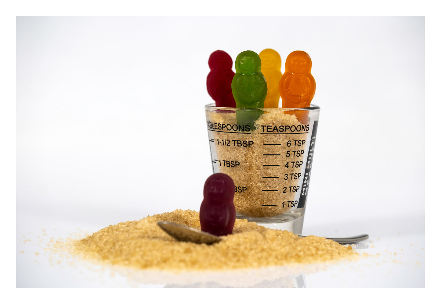 Five colourful confectionery figures arranged around a measuring glass filled with raw sugar, with a teaspoon and spilled sugar in Spoonful Of Sugar - 267000v1.