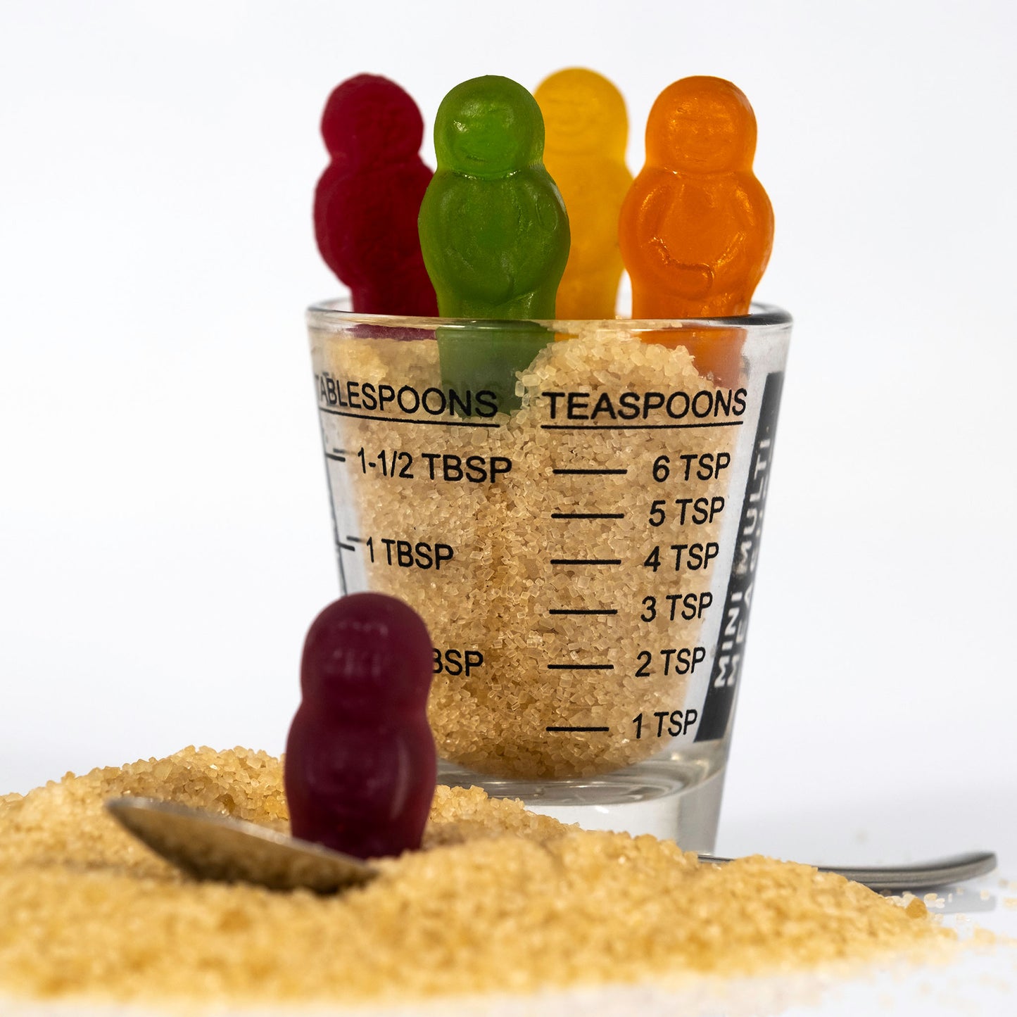 Colourful confectionery figures emerge from a measuring glass filled with raw sugar while a purple figure stands beside a teaspoon in Spoonful Of Sugar - 267000v1.