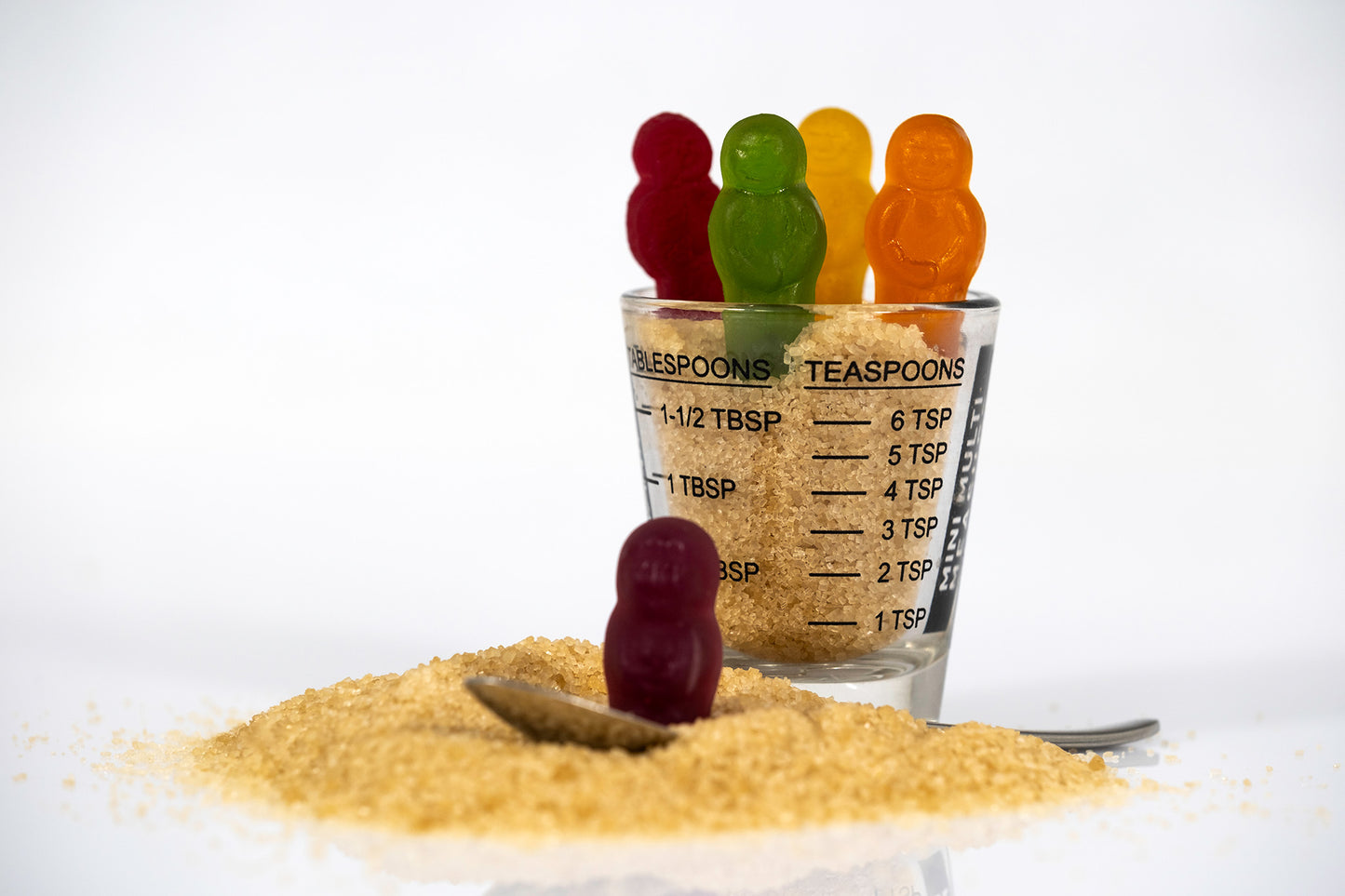 Five colourful confectionery figures gather around a sugar-filled measuring glass with a purple figure standing in spilled raw sugar beside a teaspoon.