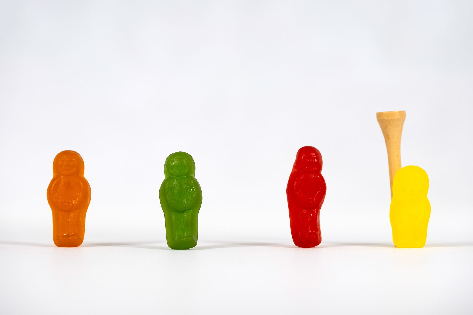 Four colourful confectionery figures stand across a minimal white landscape with a wooden golf tee positioned behind the yellow figure in Teeing Off - 267089v1.