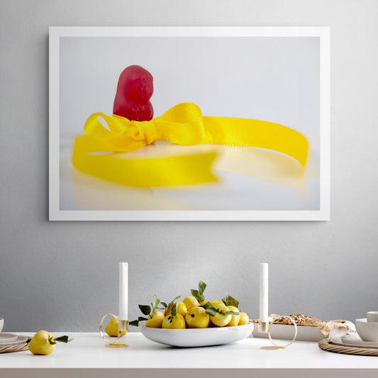 Tie A Yellow Ribbon - 267070v1 framed photographic artwork featuring a red confectionery figure behind a bright yellow satin ribbon tied in a bow, displayed in a contemporary interior.