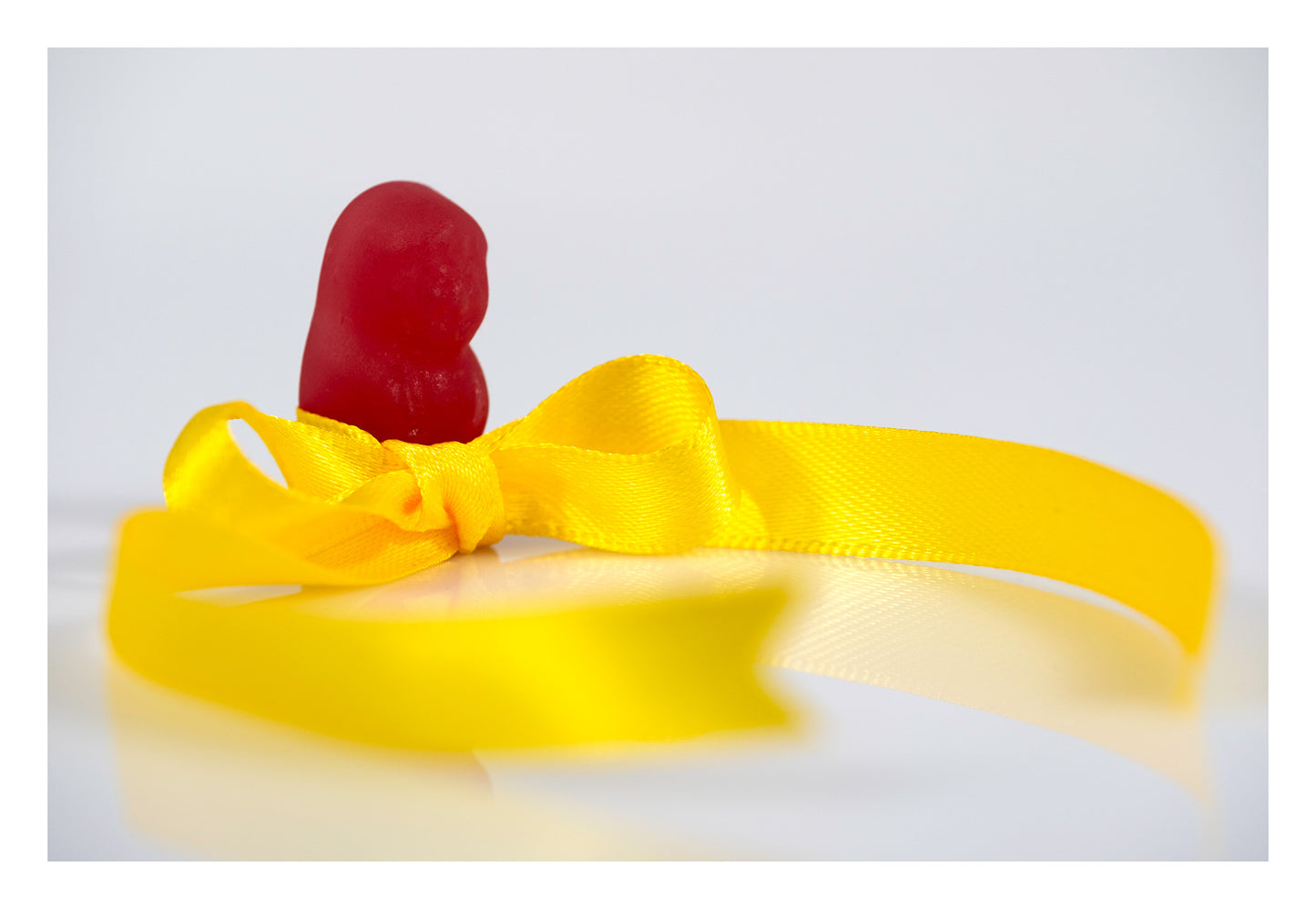 A red confectionery figure stands behind a vivid yellow satin ribbon tied into a bow against a minimal white background in Tie A Yellow Ribbon - 267070v1.