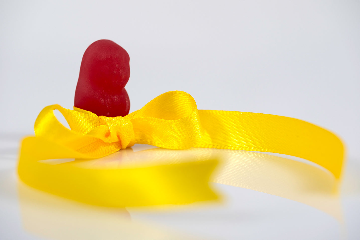 A solitary red confectionery figure stands behind a yellow satin bow as looping ribbon sweeps across a reflective white surface in Tie A Yellow Ribbon - 267070v1.
