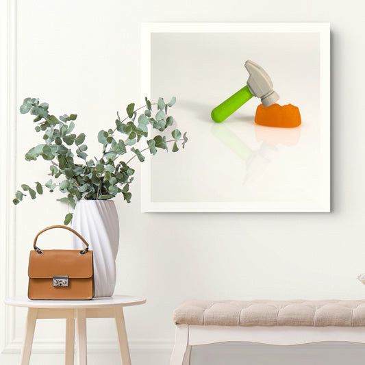 Under The Hammer - 266707v2 framed photographic artwork featuring a miniature hammer with a green handle poised above an orange confectionery form, displayed in a contemporary interior.