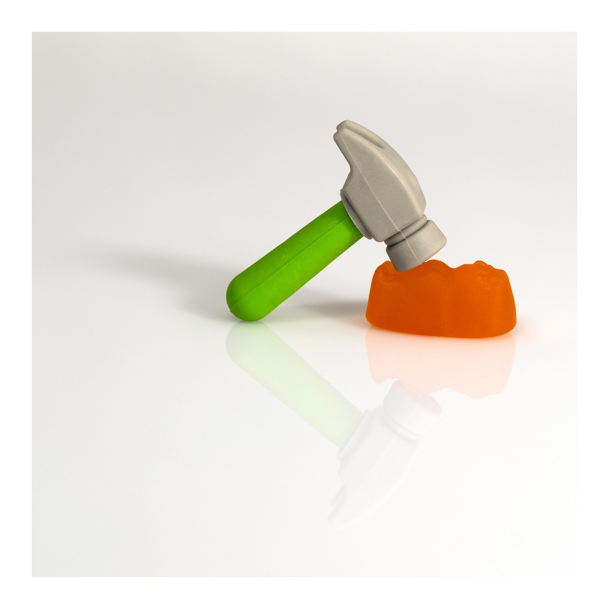 A miniature grey hammer with a vivid green handle rests above an orange confectionery form on a reflective white surface in Under The Hammer - 266707v2.