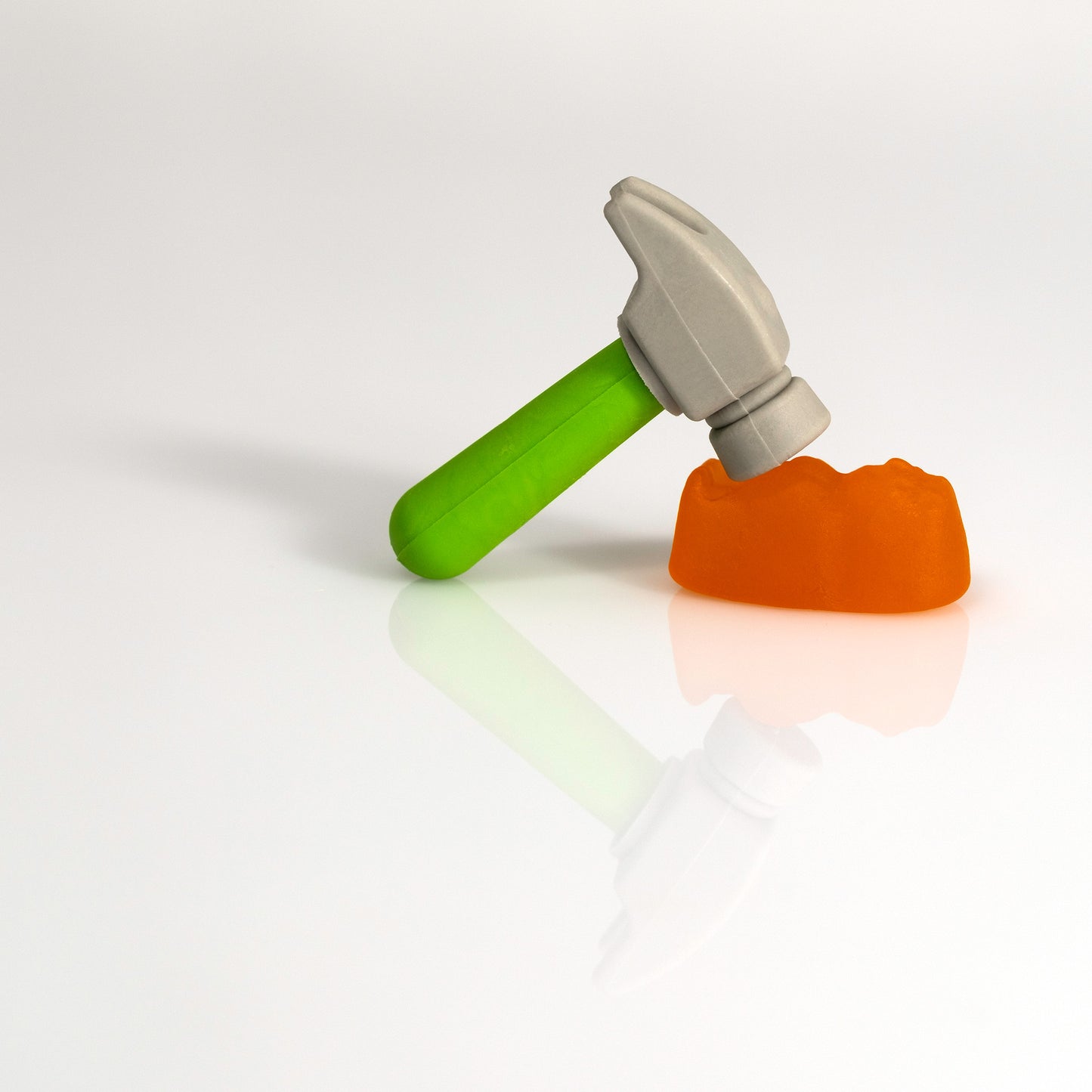 A miniature hammer with a green handle leans towards an orange confectionery form, both reflected on a minimal white surface in Under The Hammer - 266707v2.
