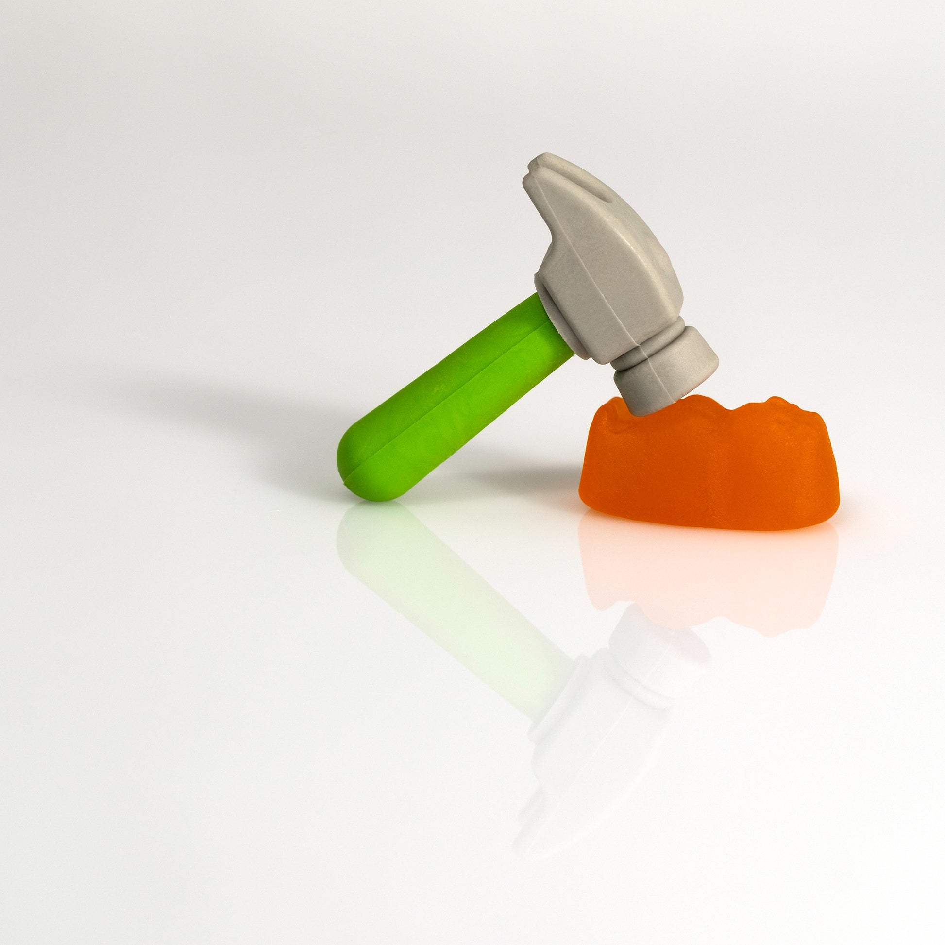 A miniature hammer with a green handle leans towards an orange confectionery form, both reflected on a minimal white surface in Under The Hammer - 266707v2.