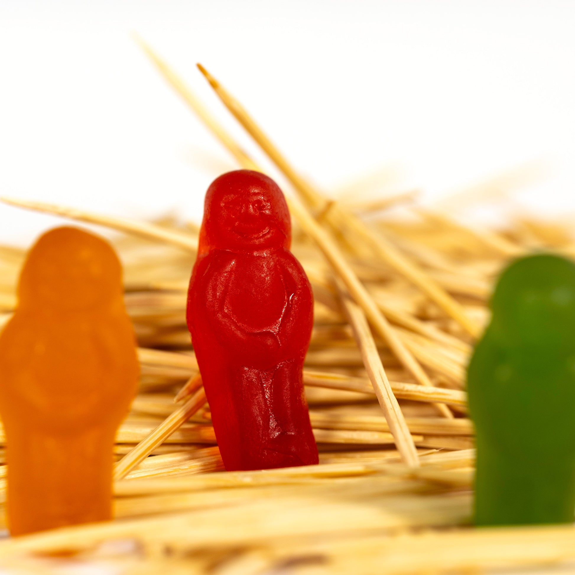 Close view of a red confectionery figure standing among intersecting wooden toothpicks, framed by softly blurred orange and green figures.