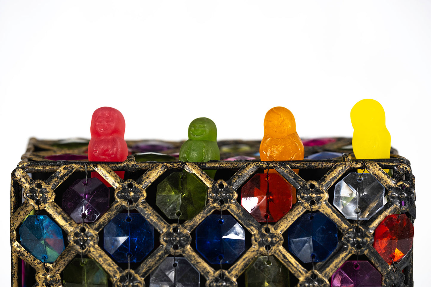 Four colourful confectionery figures arranged along the top of an ornate gold and multicoloured jewel-covered object against a white background.