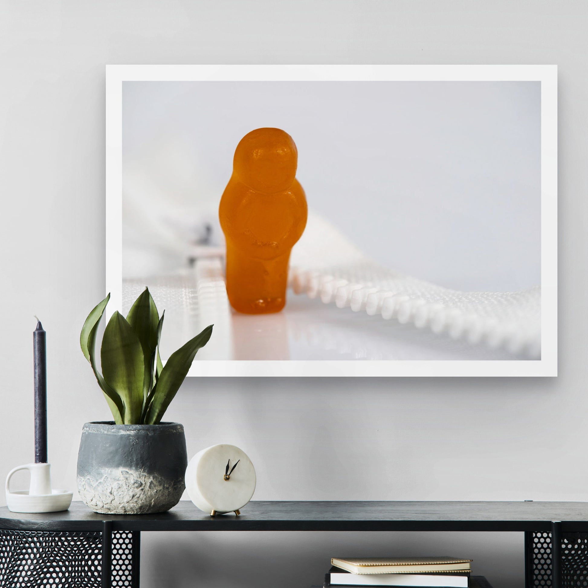 Zip Me Up - 267057v1 photographic artwork featuring an orange confectionery figure standing between the open teeth of a white zip, displayed in a contemporary interior.
