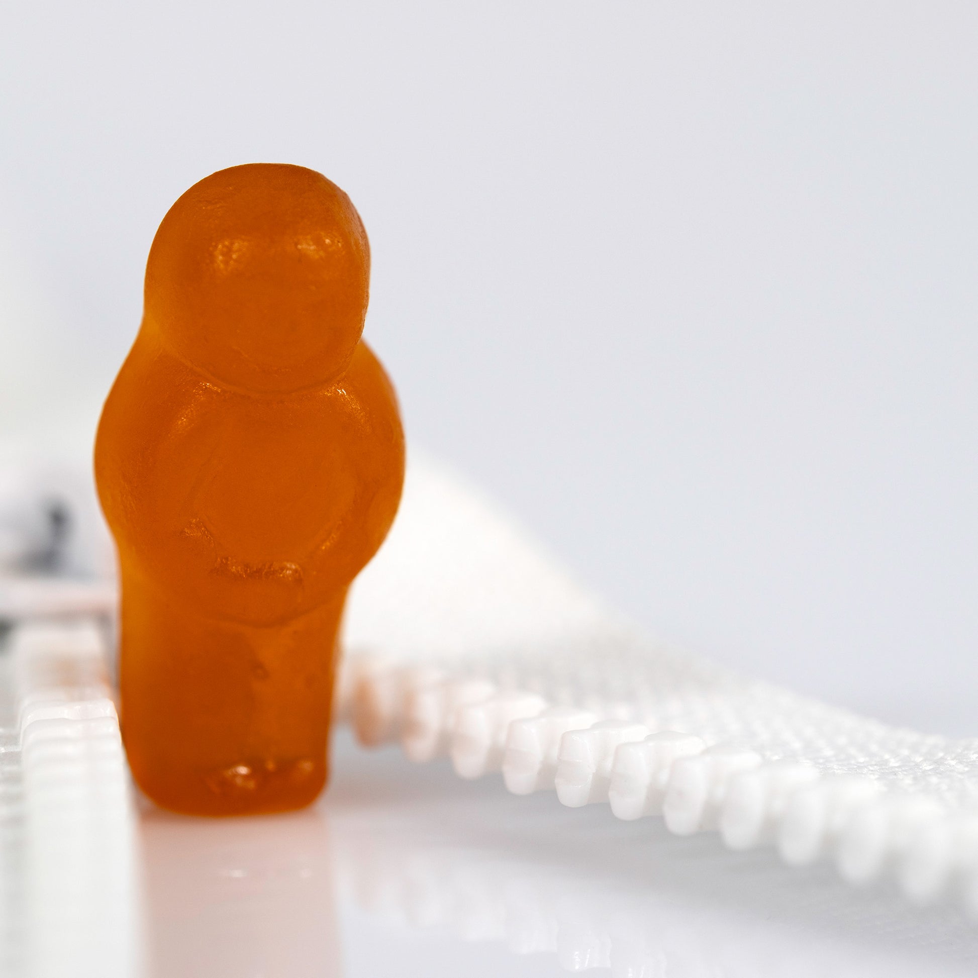 Close crop of an orange confectionery figure beside the white teeth of an open zip with expansive white negative space.