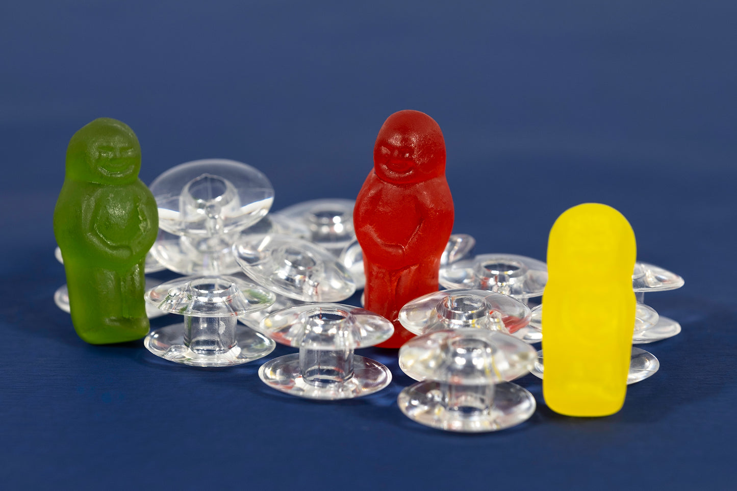Bobbin Around still life photograph of green, red and yellow jelly baby figurines arranged among transparent sewing machine bobbins on blue.
