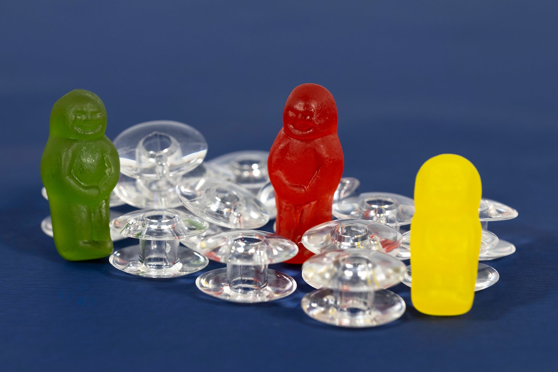 Bobbin Around still life photograph of green, red and yellow jelly baby figurines arranged among transparent sewing machine bobbins on blue.