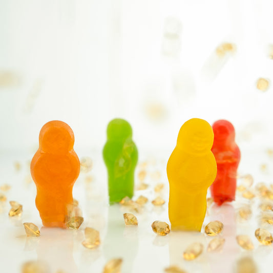All that Glitters 2871v2 - Close Detail View, colourful jelly baby figurines in orange, green, yellow, red, and purple standing upright among scattered golden crystal fragments on a glossy white surface; playful, pop-art style fine art print by Raymond Mather, ART1.